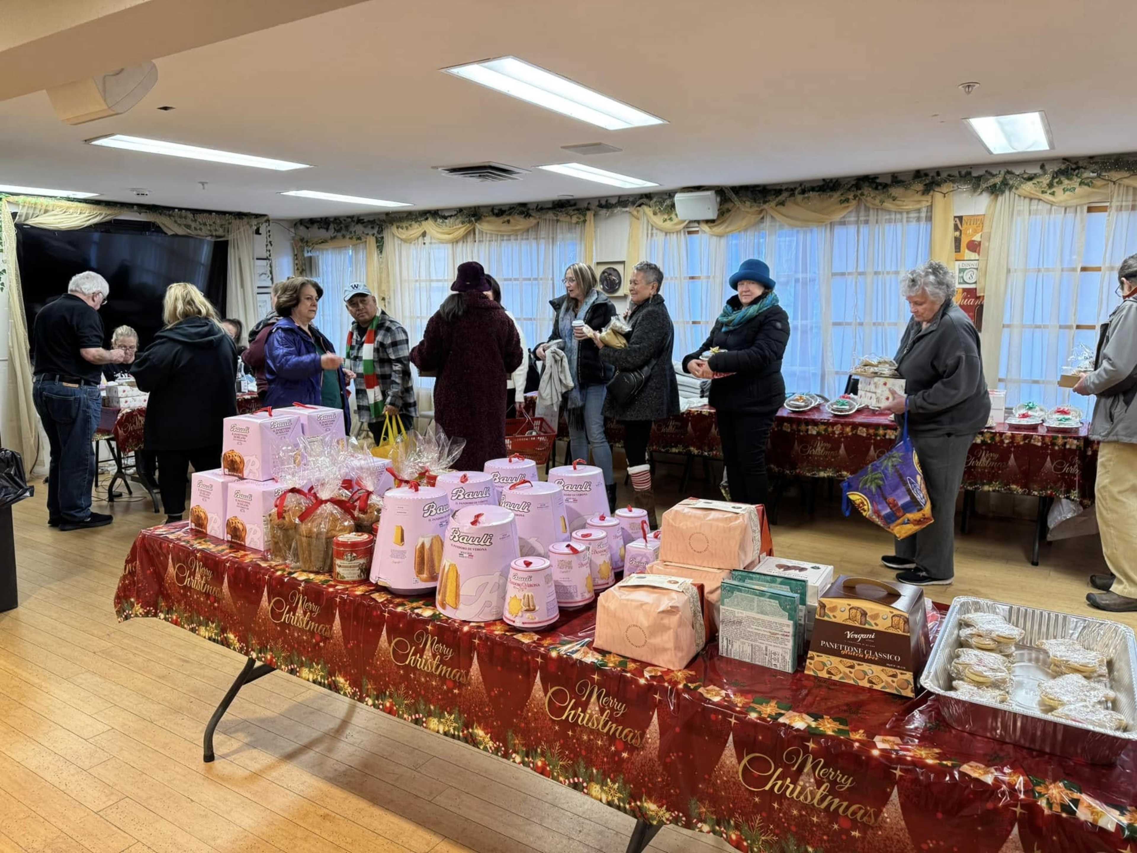 A group of people gathers around a table covered with various holiday-themed baked goods and treats in a festively decorated room.
