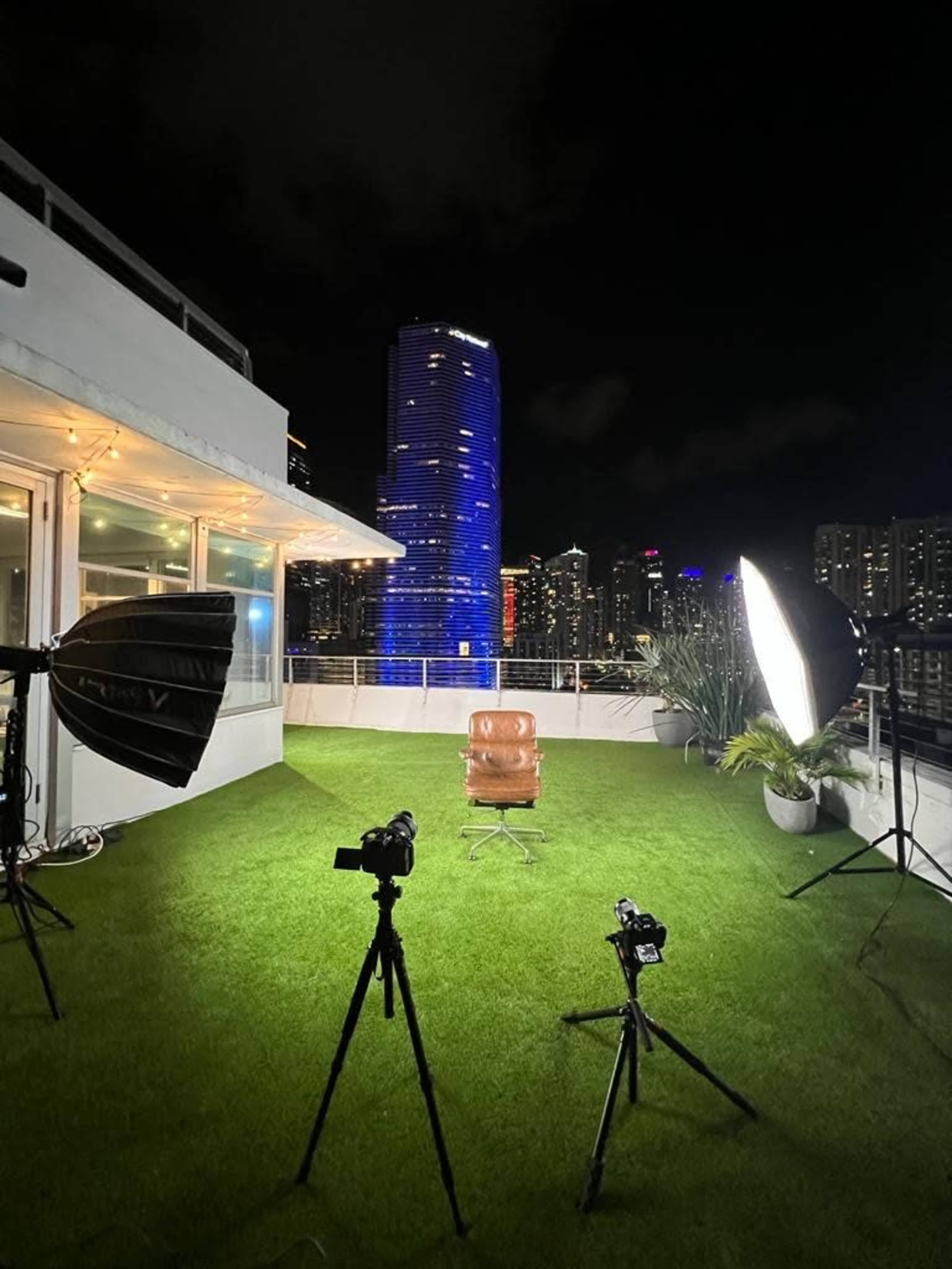 A rooftop setting at night features a brown leather chair, surrounded by three cameras and bright studio lights, with a city skyline illuminated in the background.