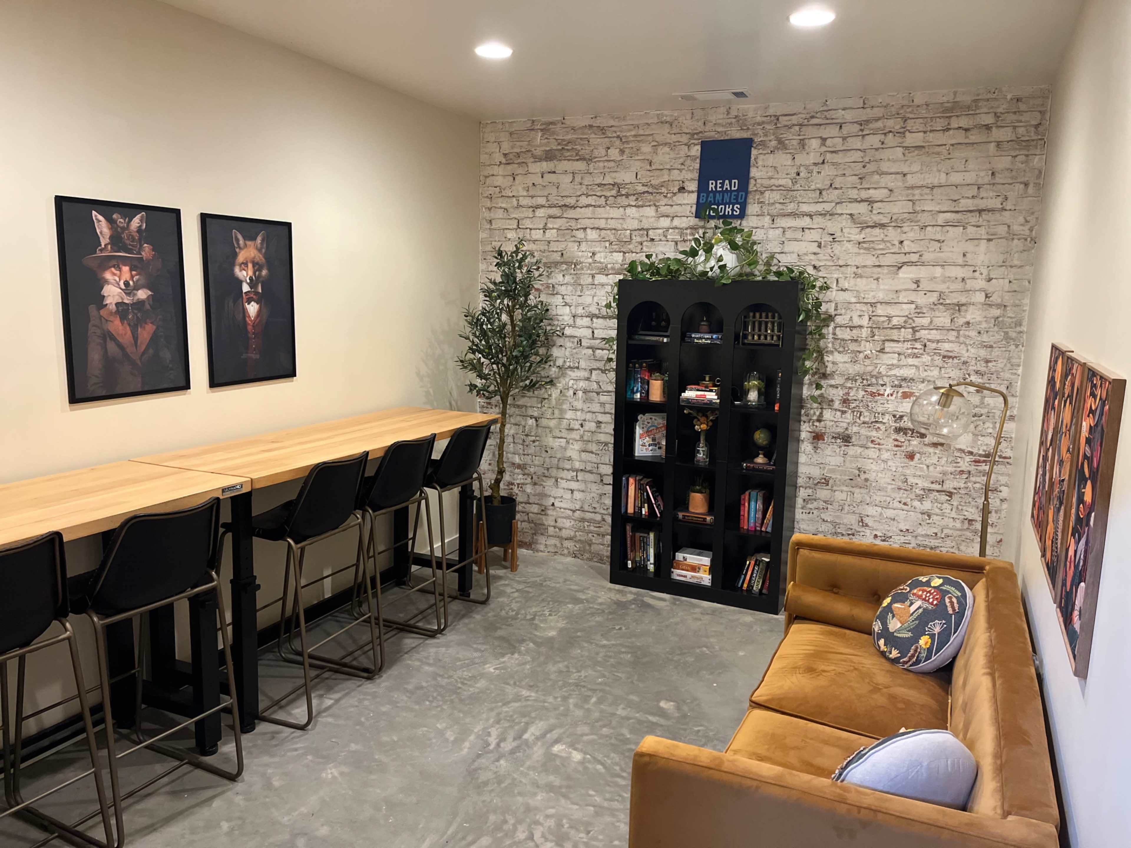 The image shows a modern room featuring three framed animal portraits on the wall, a long table with high chairs, a black bookshelf filled with books, and a brown couch against a brick wall.