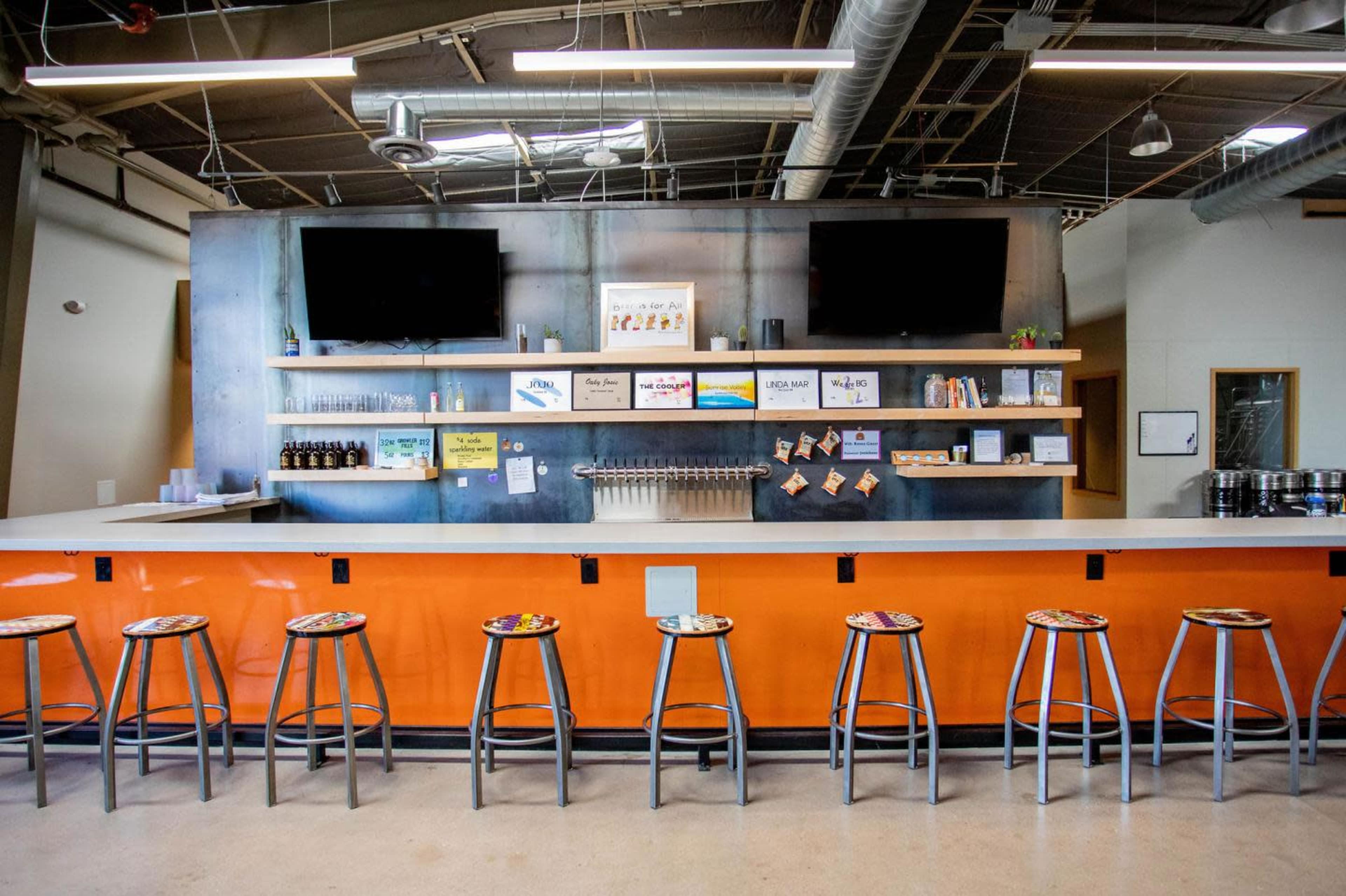 The image shows a sleek bar area with a white countertop, orange accents, and several high stools arranged in front of it, featuring two mounted TVs and various menus and items displayed on the wall.