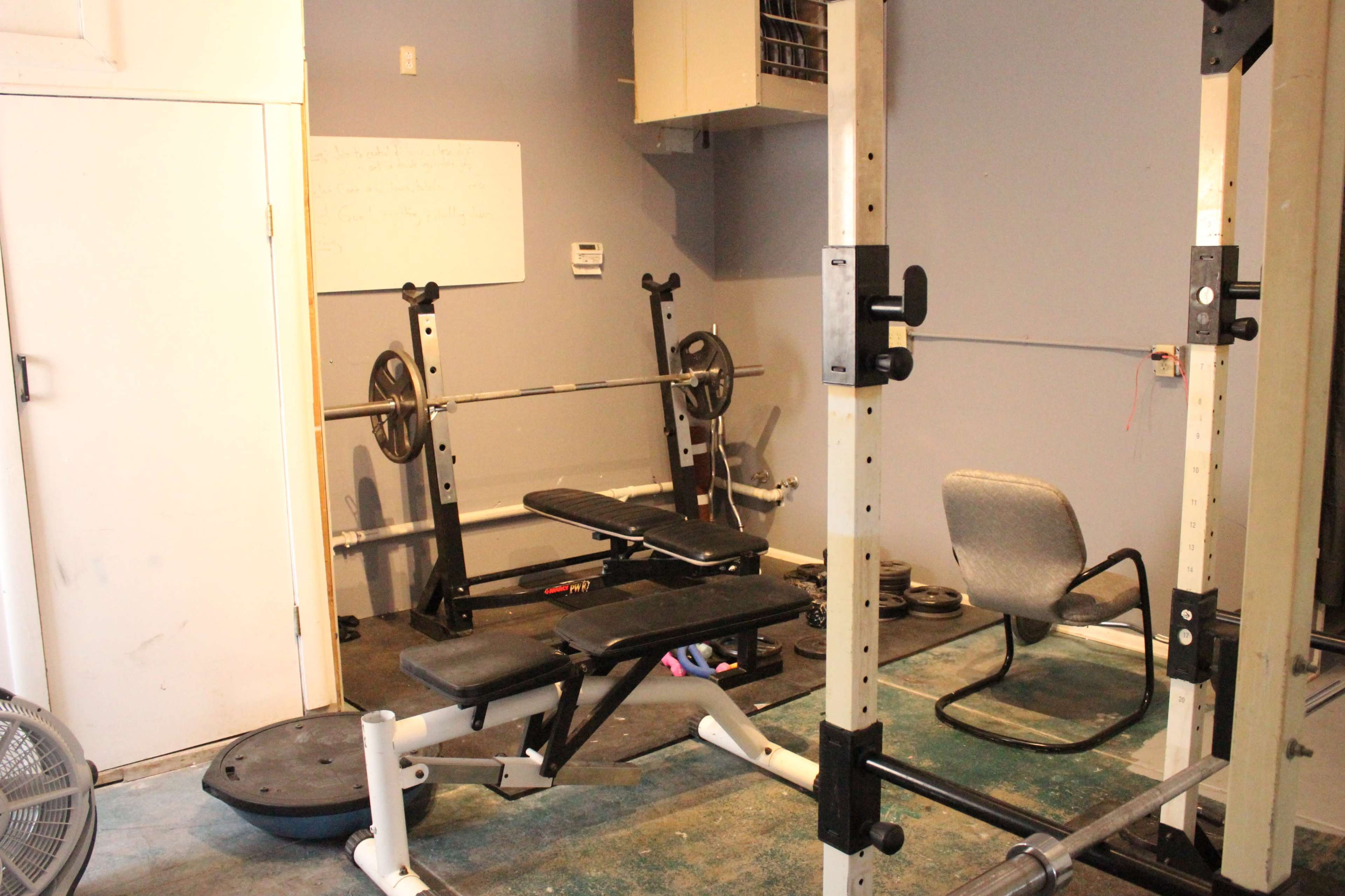 The image shows a small gym area with a weight bench, barbell rack, and a chair against a wall.