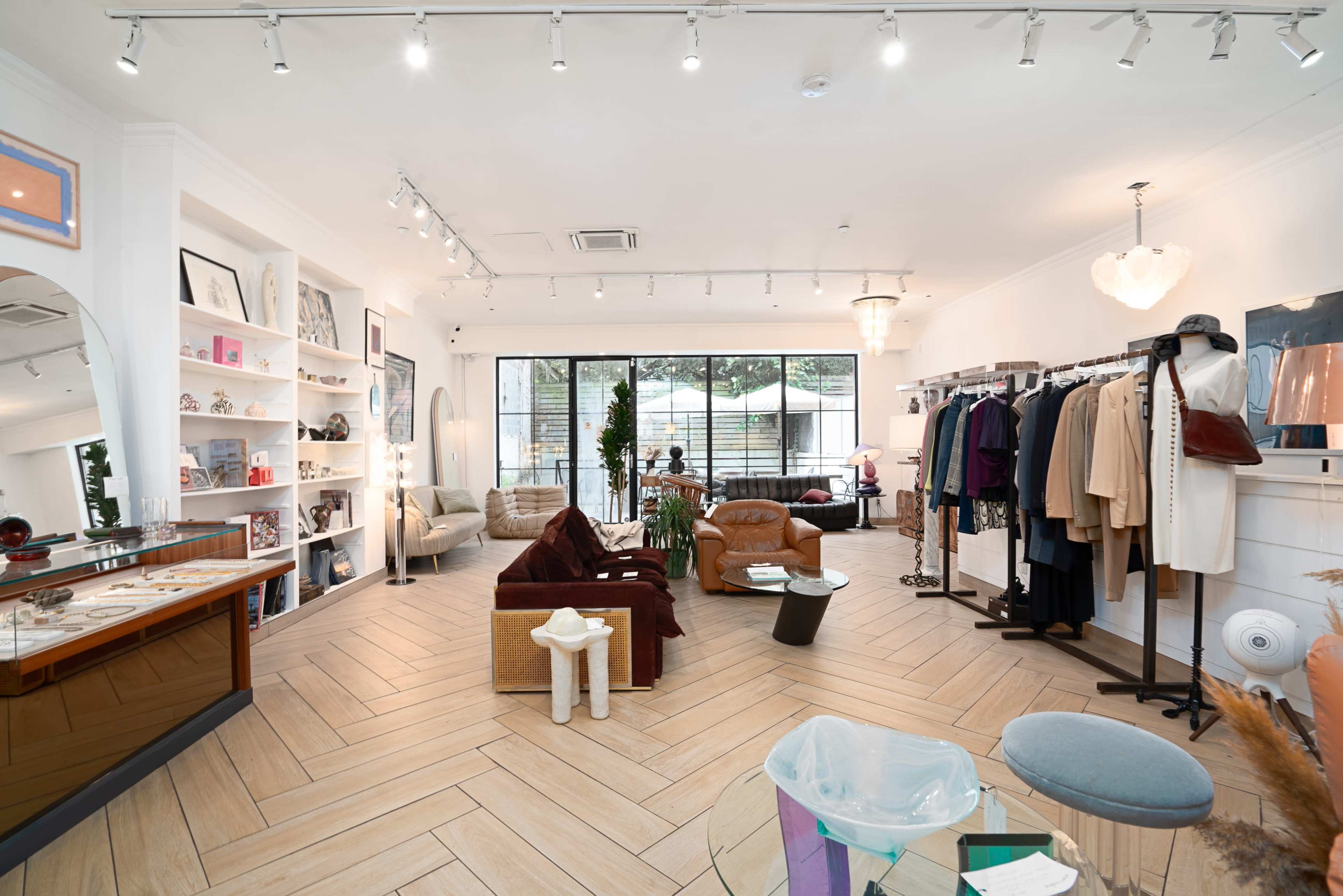 The image shows a modern clothing boutique with a mix of apparel displayed on racks, stylish seating areas, and art pieces arranged on shelves.