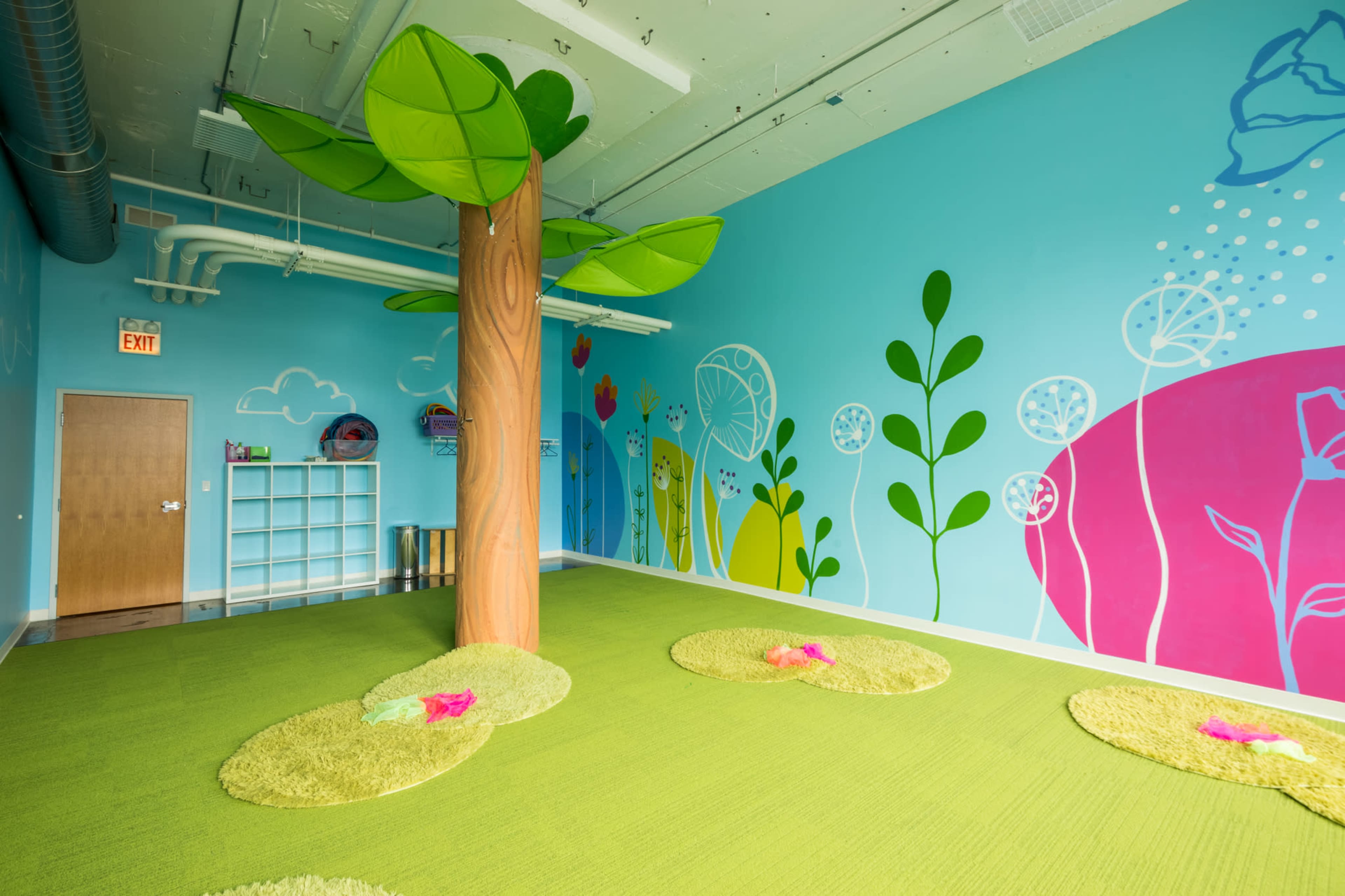 A brightly colored children's playroom with a large tree sculpture, green carpet, and whimsical wall decorations featuring flowers and plants.