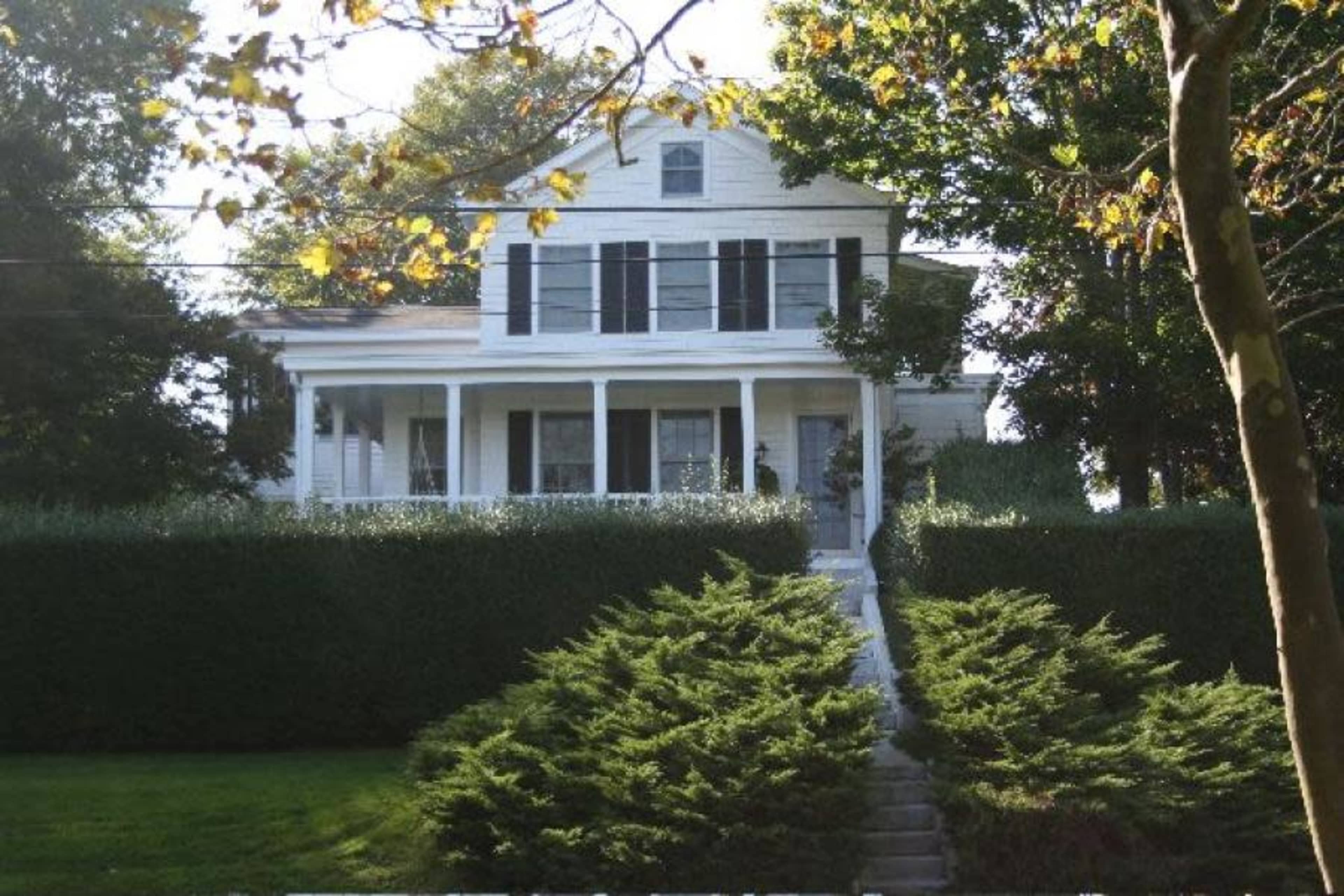 A white two-story house with black shutters is set behind a neatly trimmed hedge and a gravel pathway.