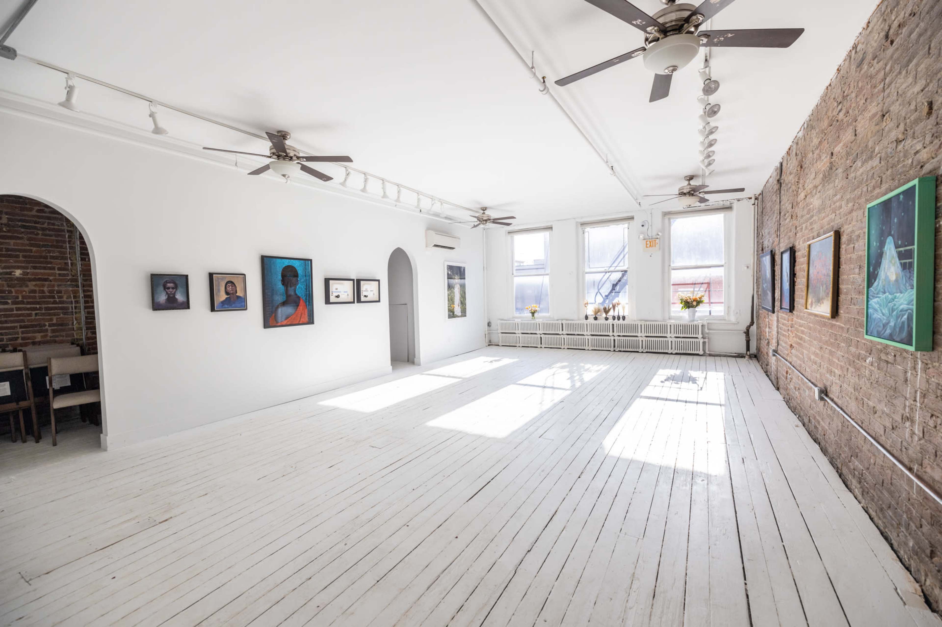 A bright art gallery featuring a white wooden floor, exposed brick wall, and several framed paintings displayed on the walls.