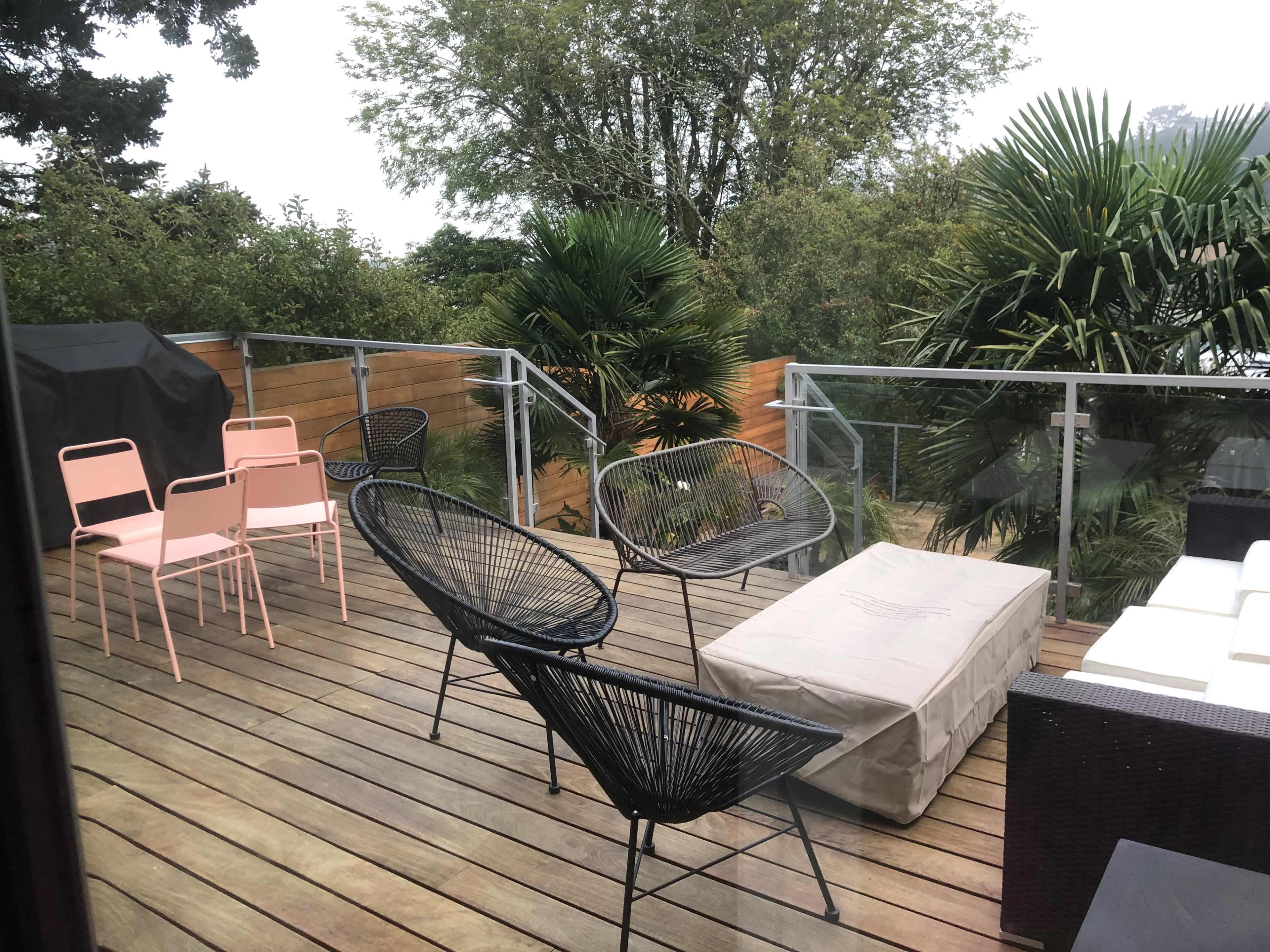 A wooden deck features a variety of outdoor furniture, including black and pink chairs, a coffee table, and tropical plants in the background.