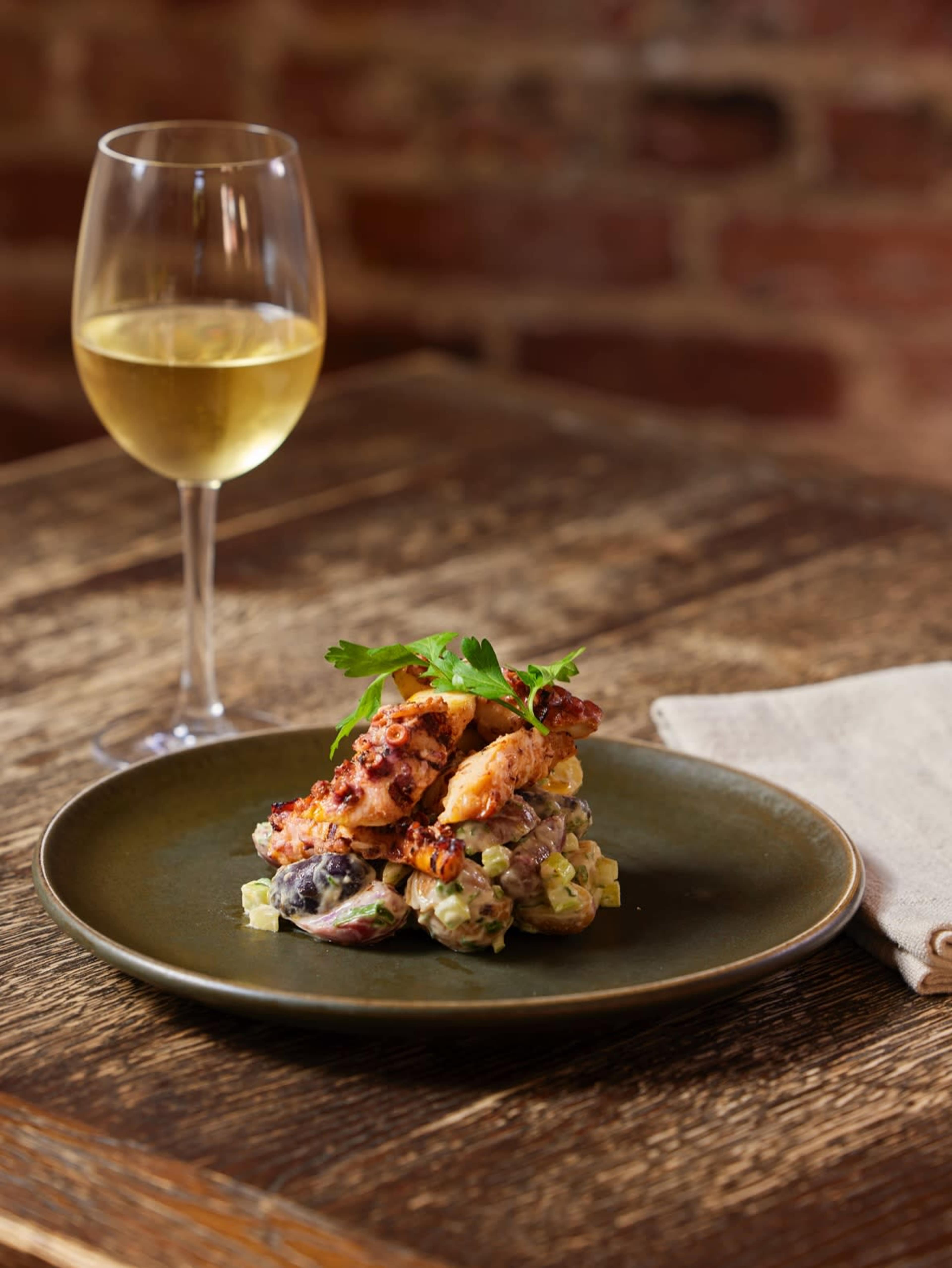 A plate of fried chicken is served atop a creamy salad with vegetables, accompanied by a glass of white wine on a wooden table.
