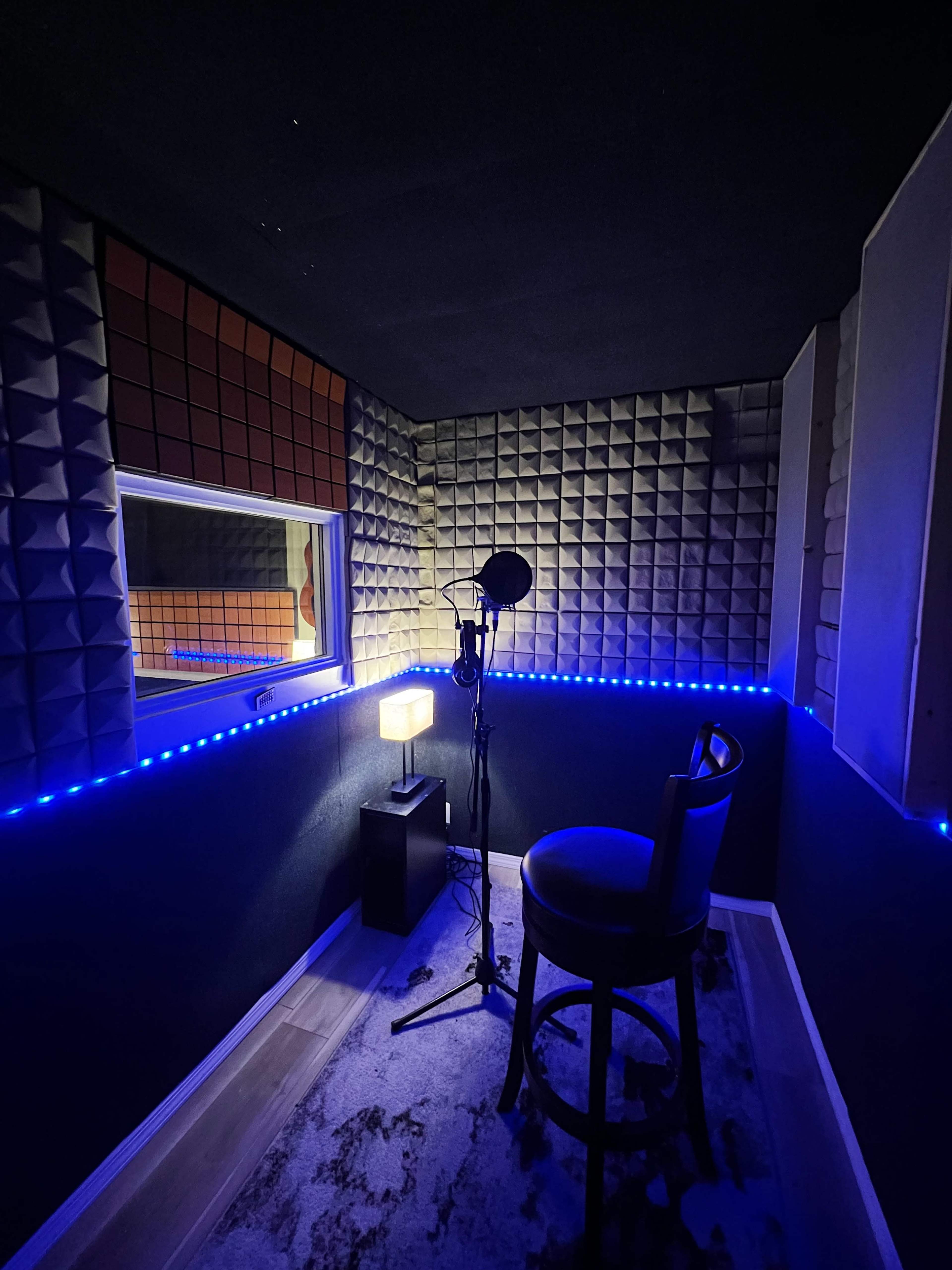 A small recording studio with soundproof walls, a microphone on a stand, a chair, and mood lighting along the edges.