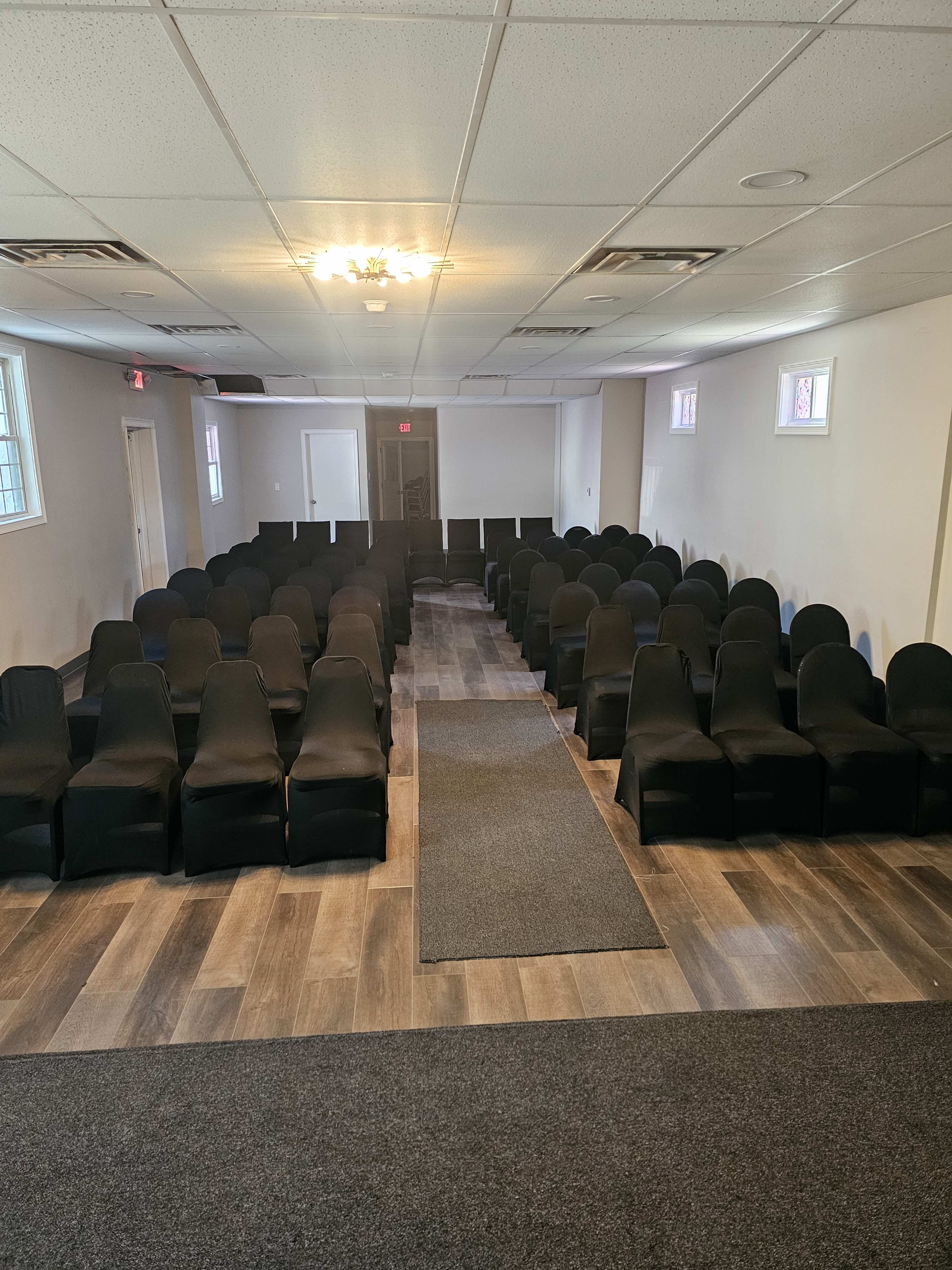 The image shows an empty room arranged with rows of black chairs and a carpeted aisle leading to a door at the back.