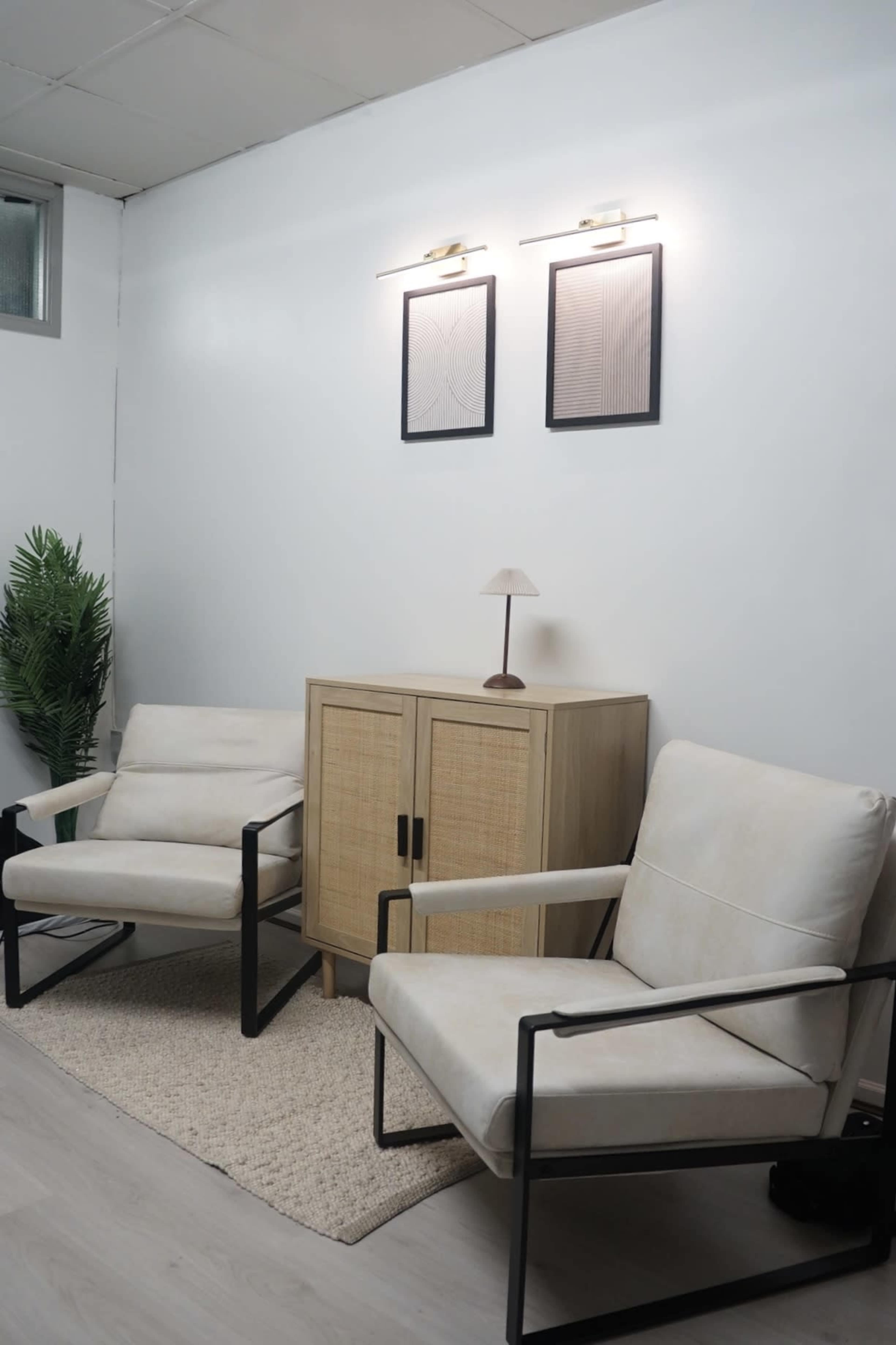 The image shows a living area featuring two armchairs, a cabinet, and wall-mounted artwork, with a plant in the corner.