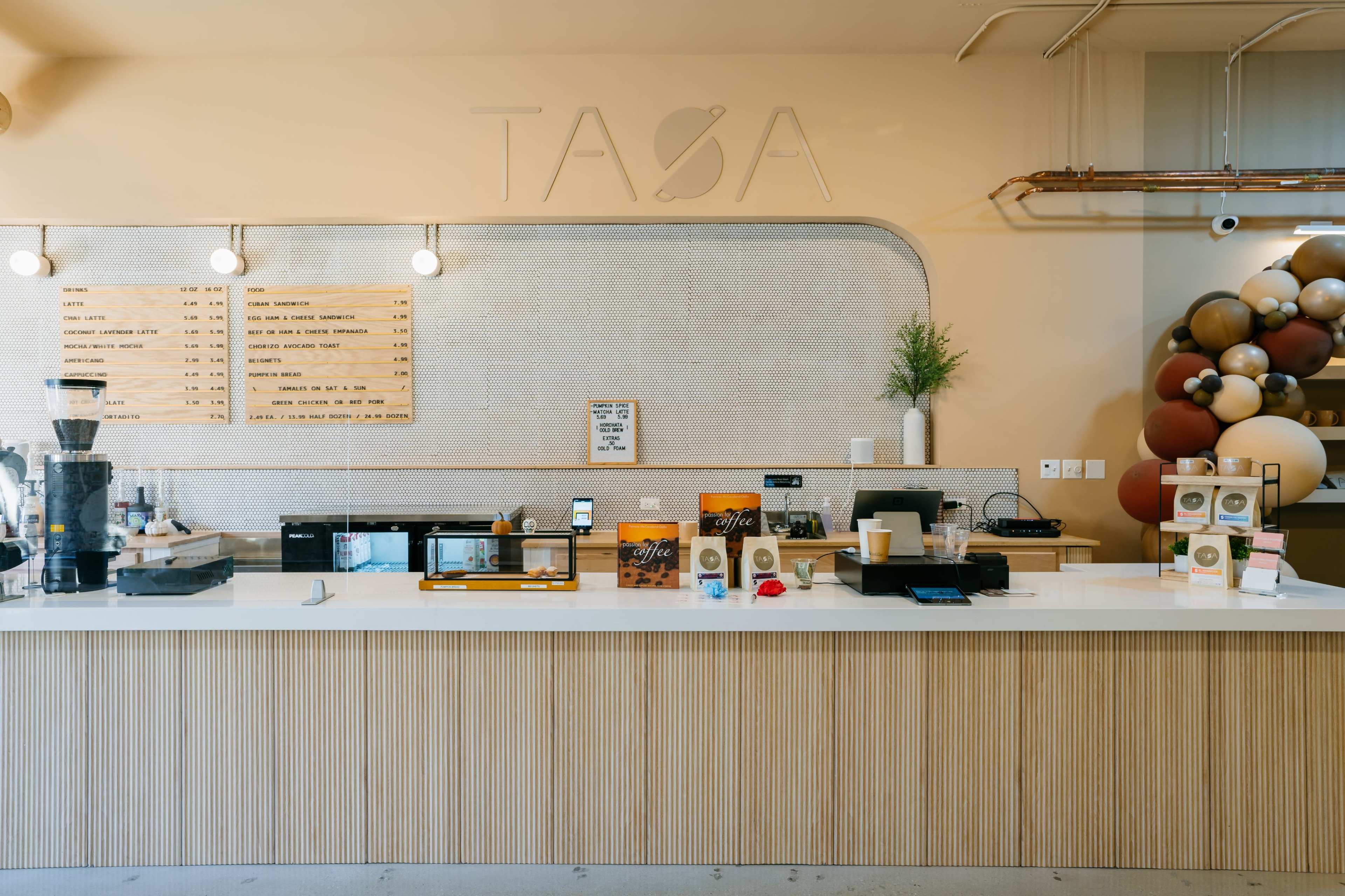 The image shows a modern coffee shop counter with a light-colored wooden facade, various menu boards, and decorative elements including a display of coffee products and a plant.