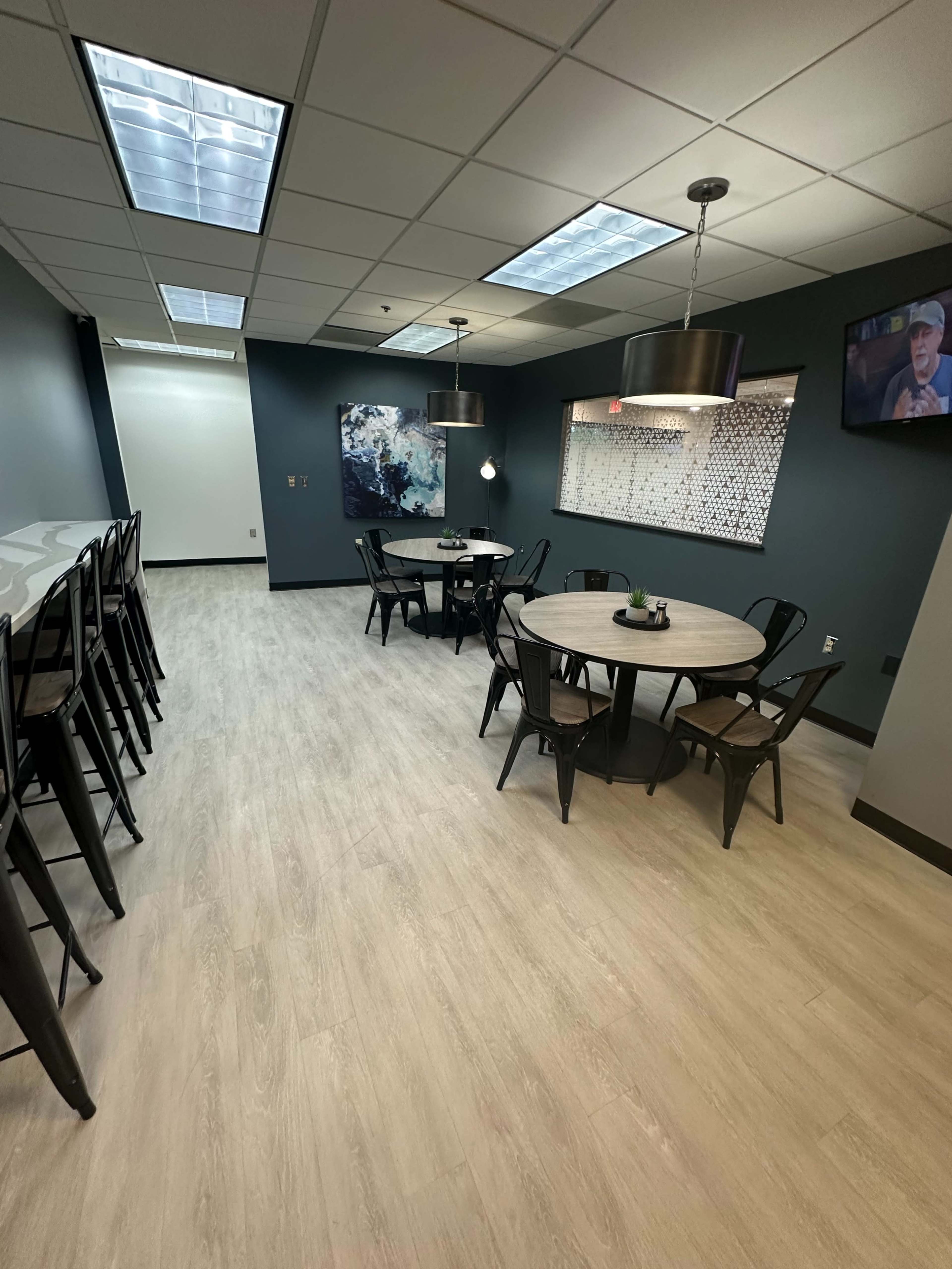A modern break room with several tables, chairs, a wall-mounted television, and a decorative wall feature, all in a well-lit, minimalistic space.