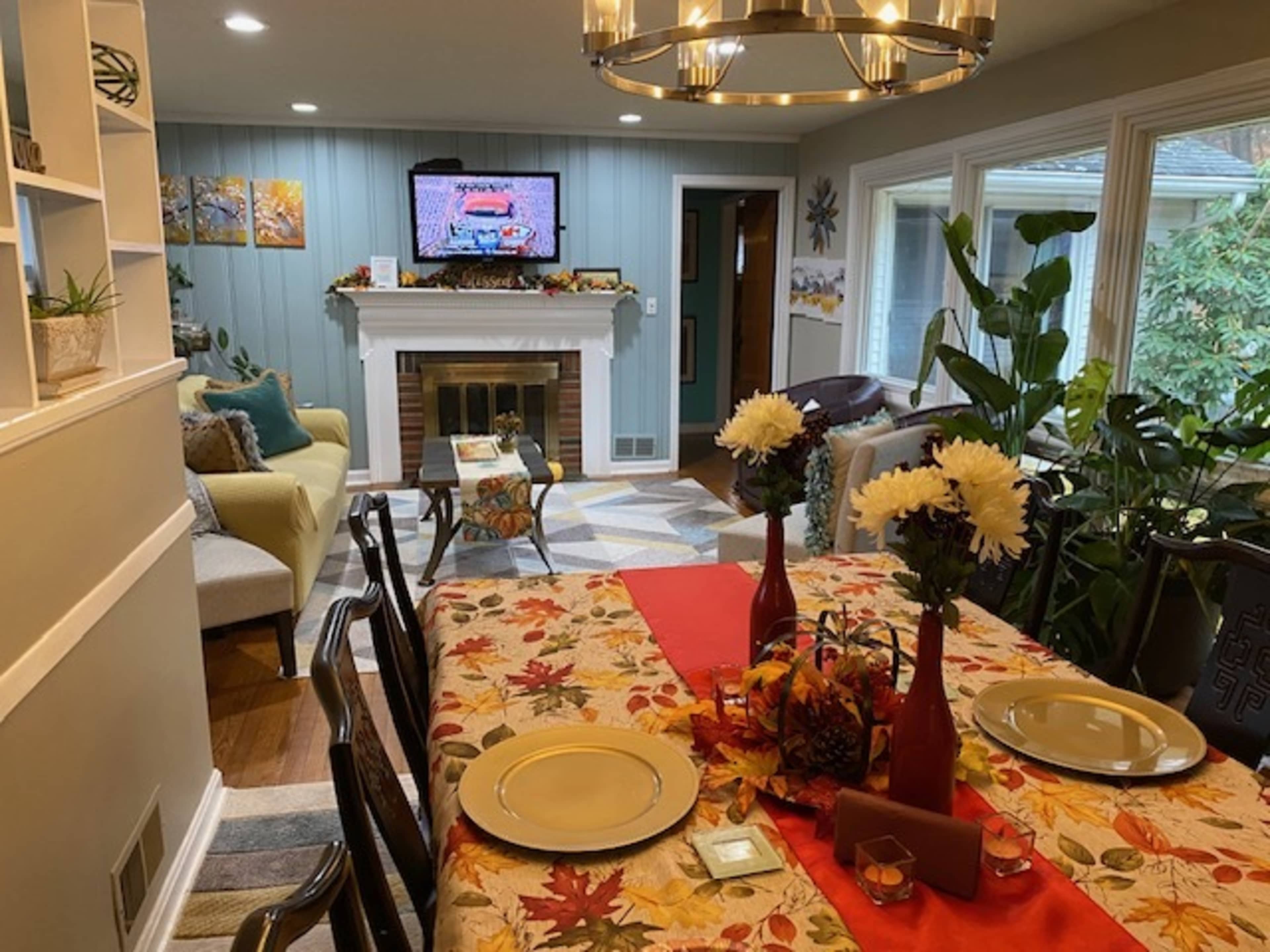 A dining area is set for a meal with a table covered in a fall-themed tablecloth and decorative elements, while a living room can be seen in the background with a television displaying a program.