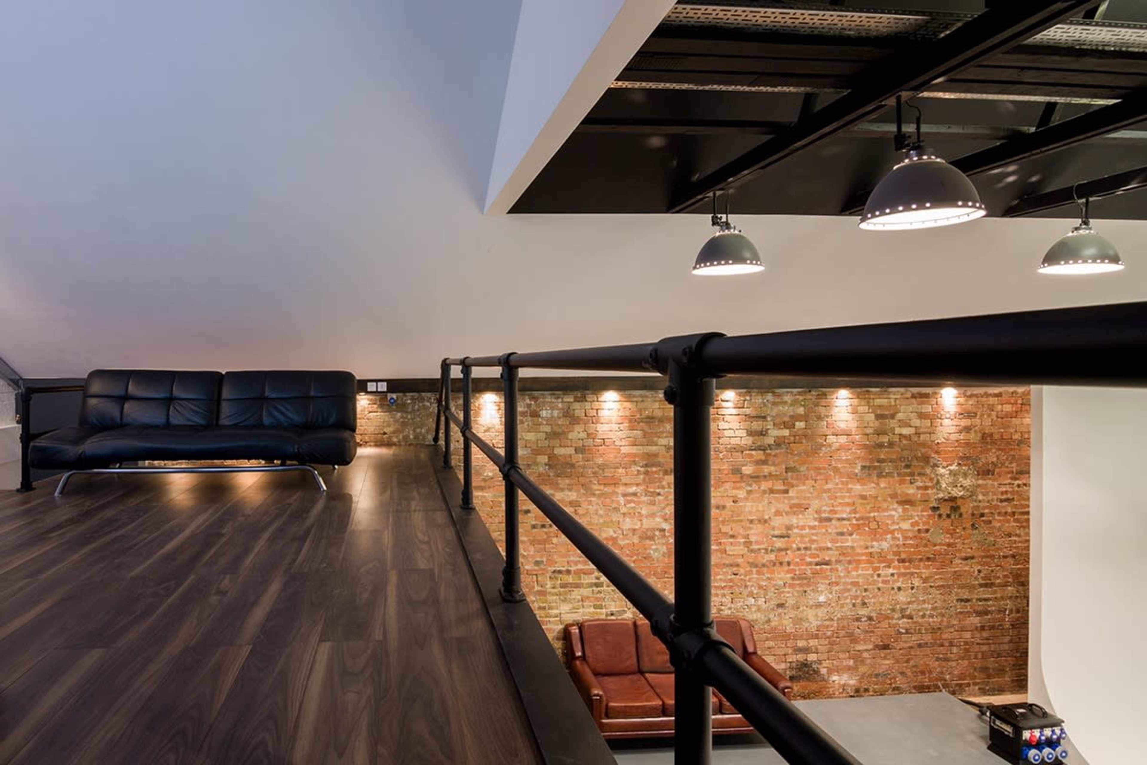 The image shows a modern loft space with a balcony overlooking a living area featuring a brick wall and two sofas.