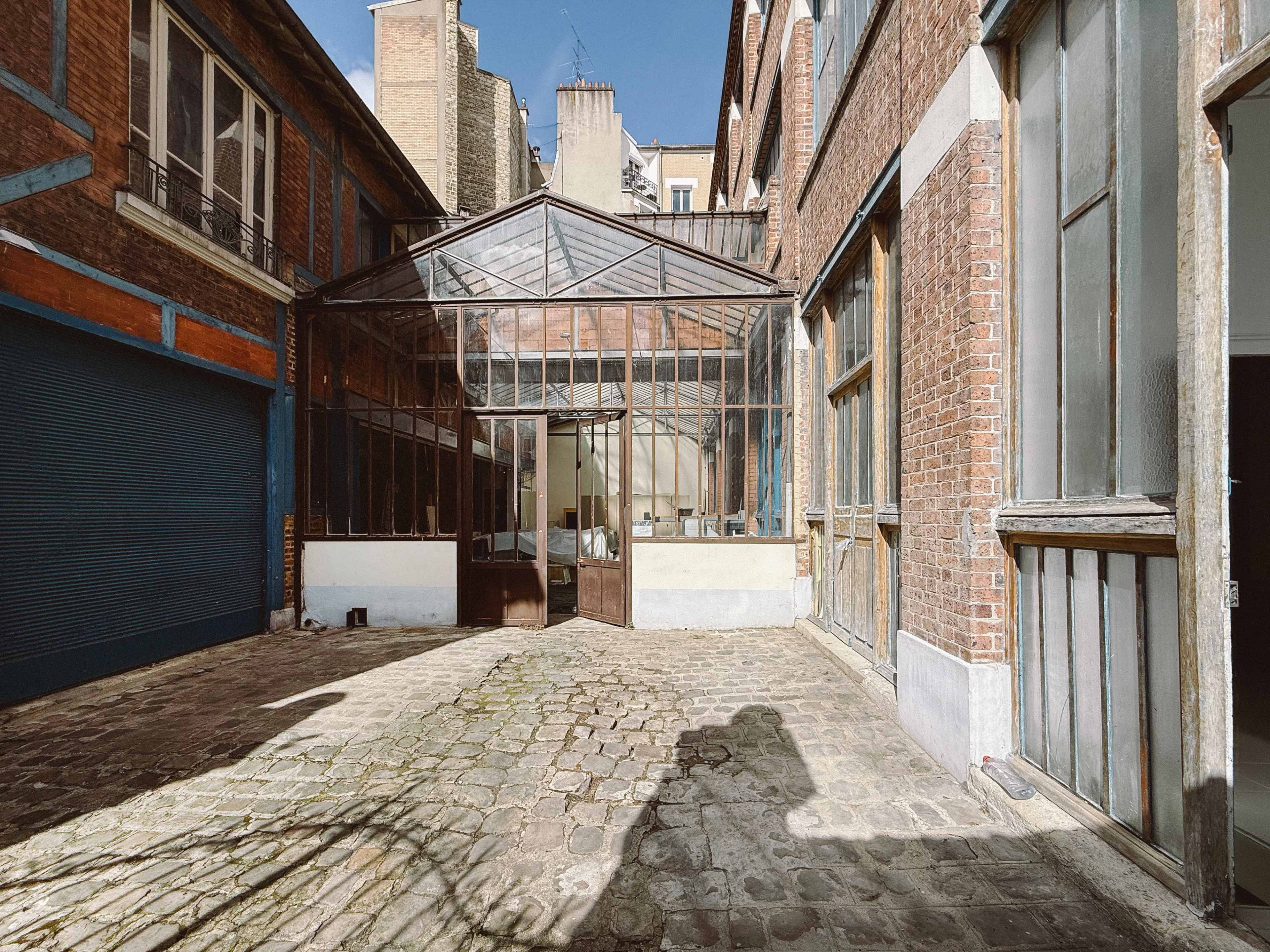 The image shows a cobblestone courtyard featuring a glass greenhouse surrounded by brick buildings.