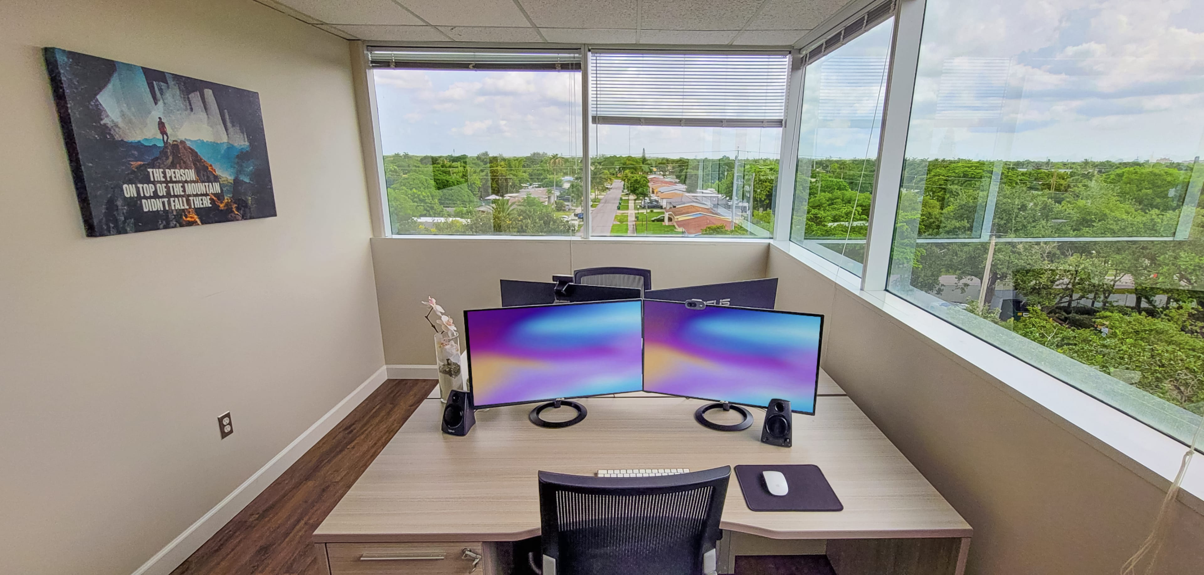 A desk with dual monitors is positioned near a large window overlooking a green landscape and road.