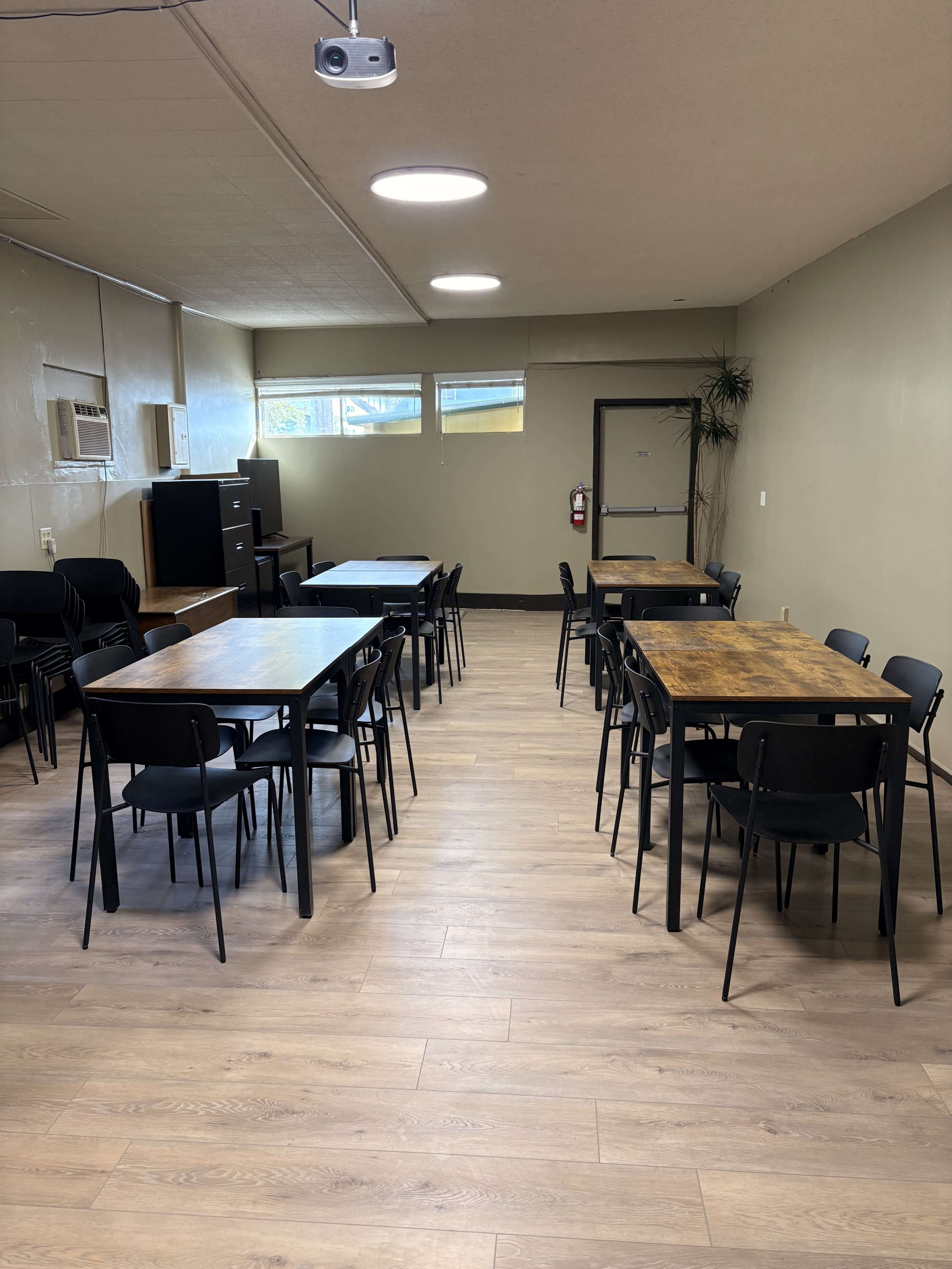 The image shows a spacious room with several tables and chairs arranged in two separate groups, along with a projector on the ceiling and a door leading to another area.