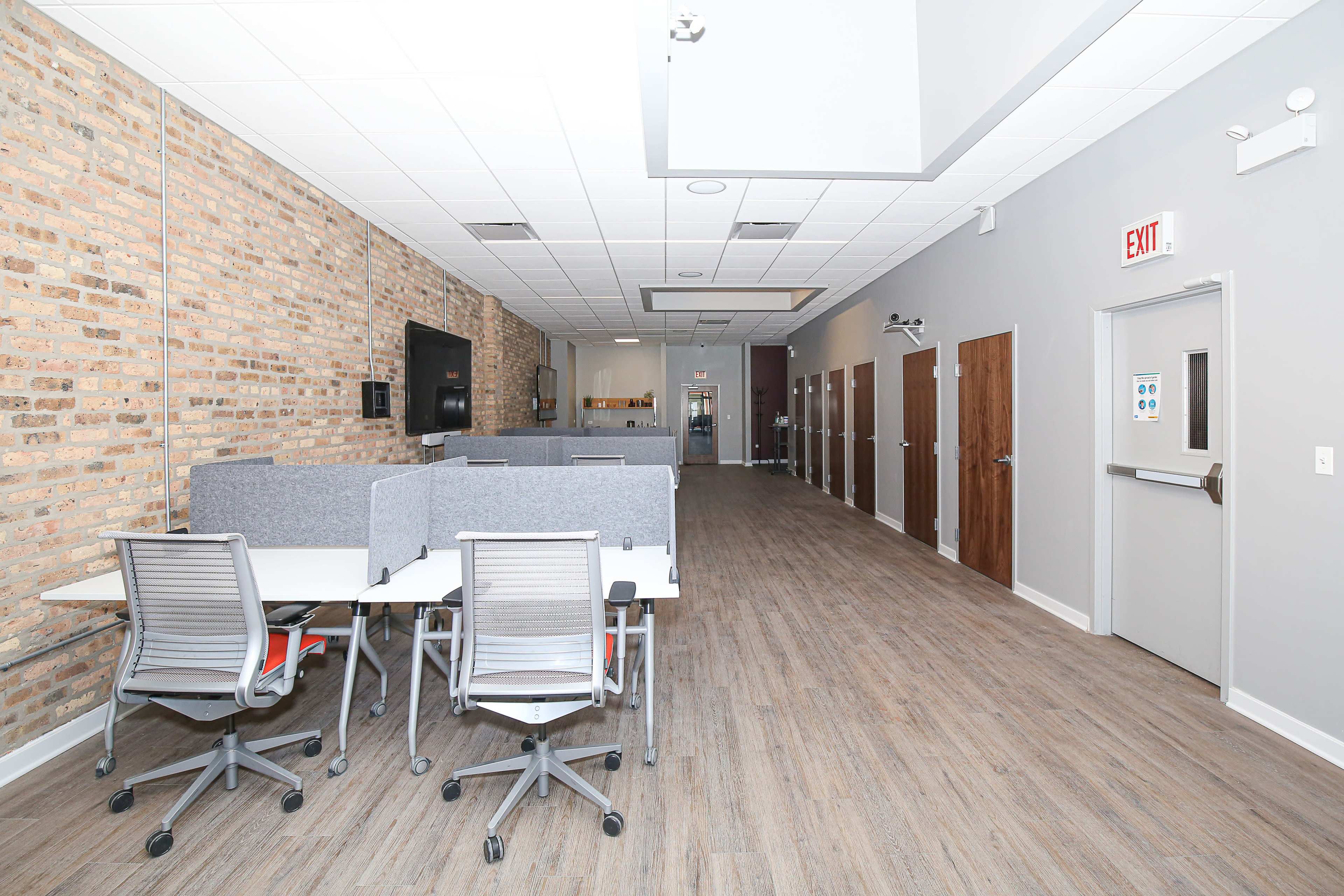 A modern office space features a brick wall, workstations with rolling chairs, and several closed doors along a corridor.