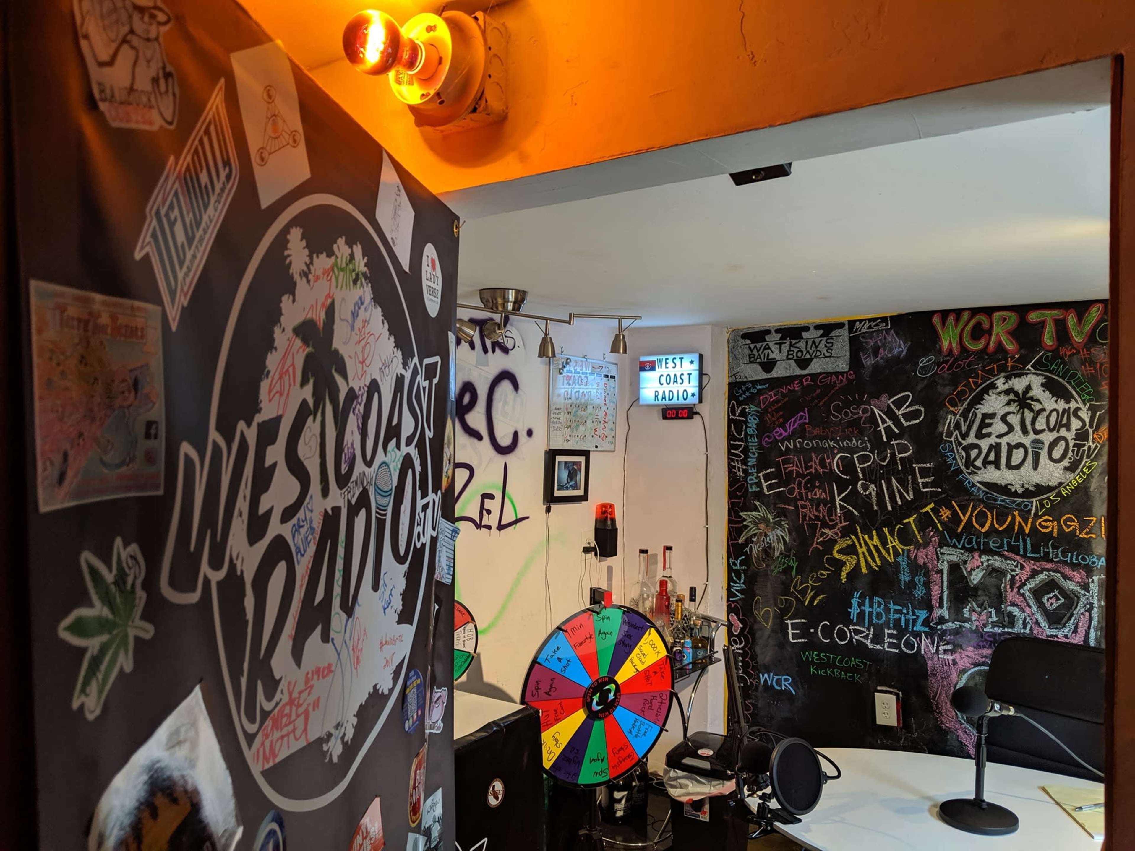 A colorful room decorated with various posters and graffiti, featuring a "West Coast Radio" sign and a prize wheel.