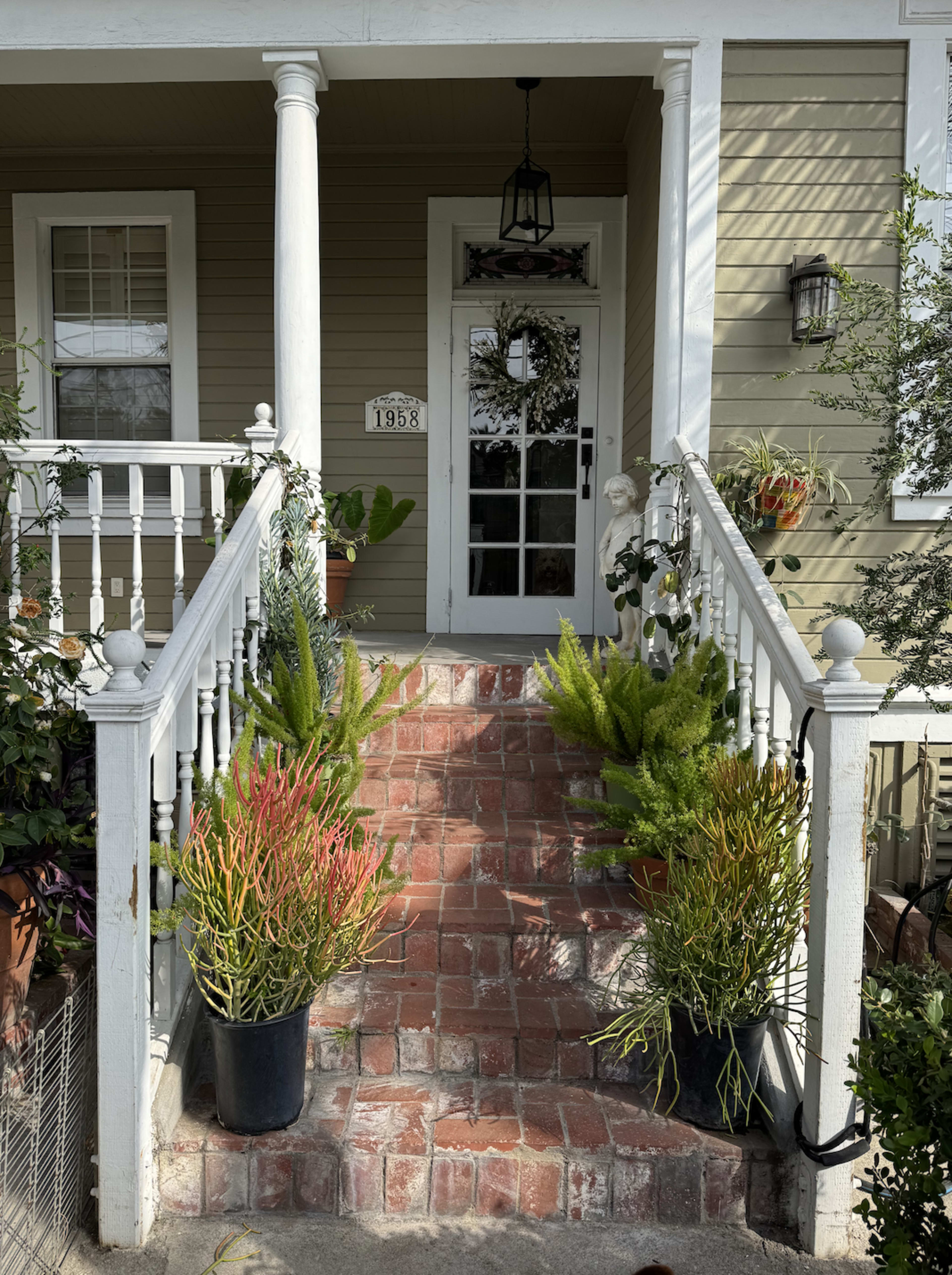 The image shows a house entrance with a brick walkway leading up to a door, flanked by potted plants on either side.