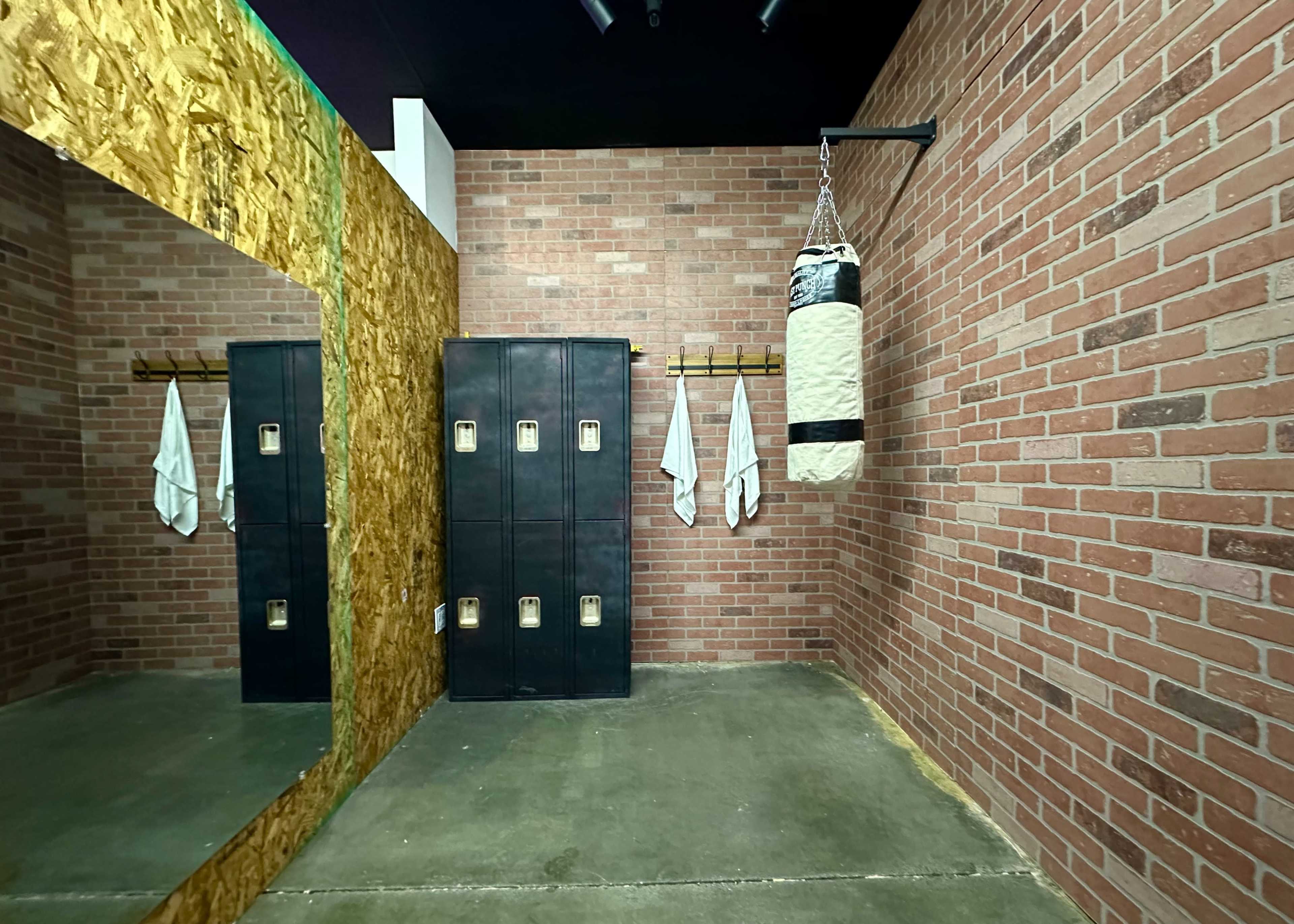 A boxing gym interior with a mirrored wall, a punching bag hanging, lockers, and towels arranged on hooks.