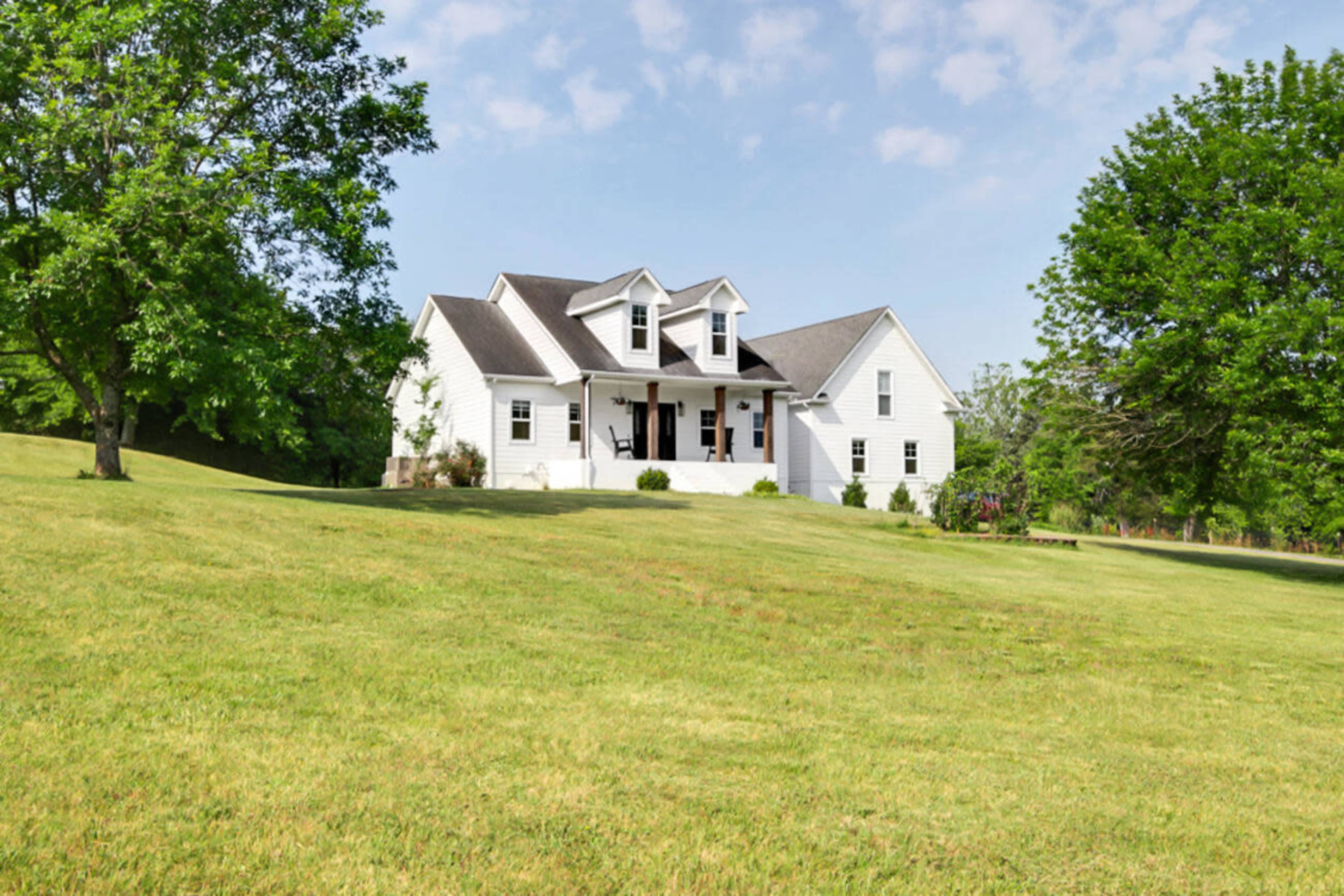 A white house with a gabled roof is situated on a grassy lawn surrounded by trees.