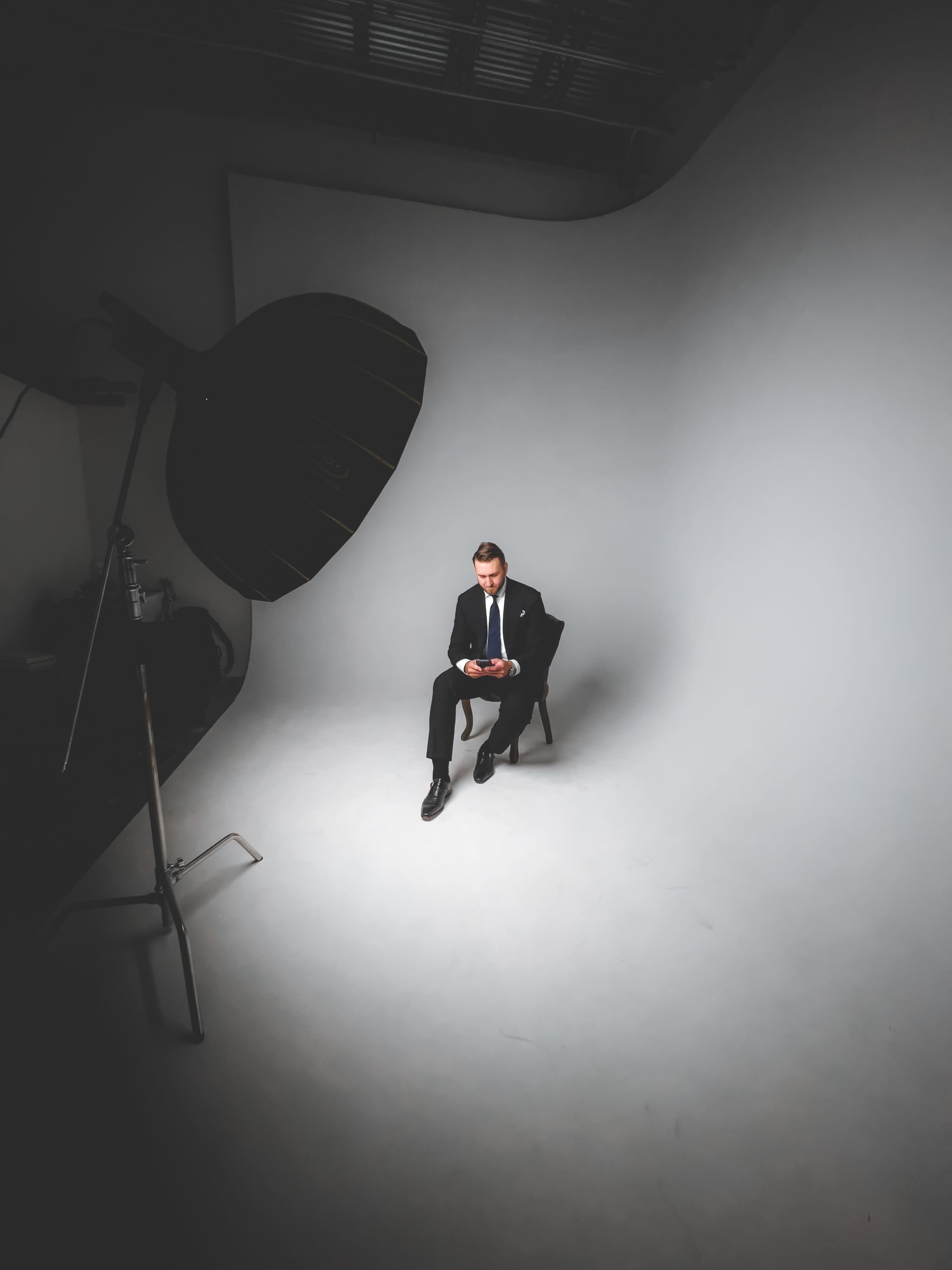 A man in a suit sits on a chair, looking at his phone in a minimalist studio setting.