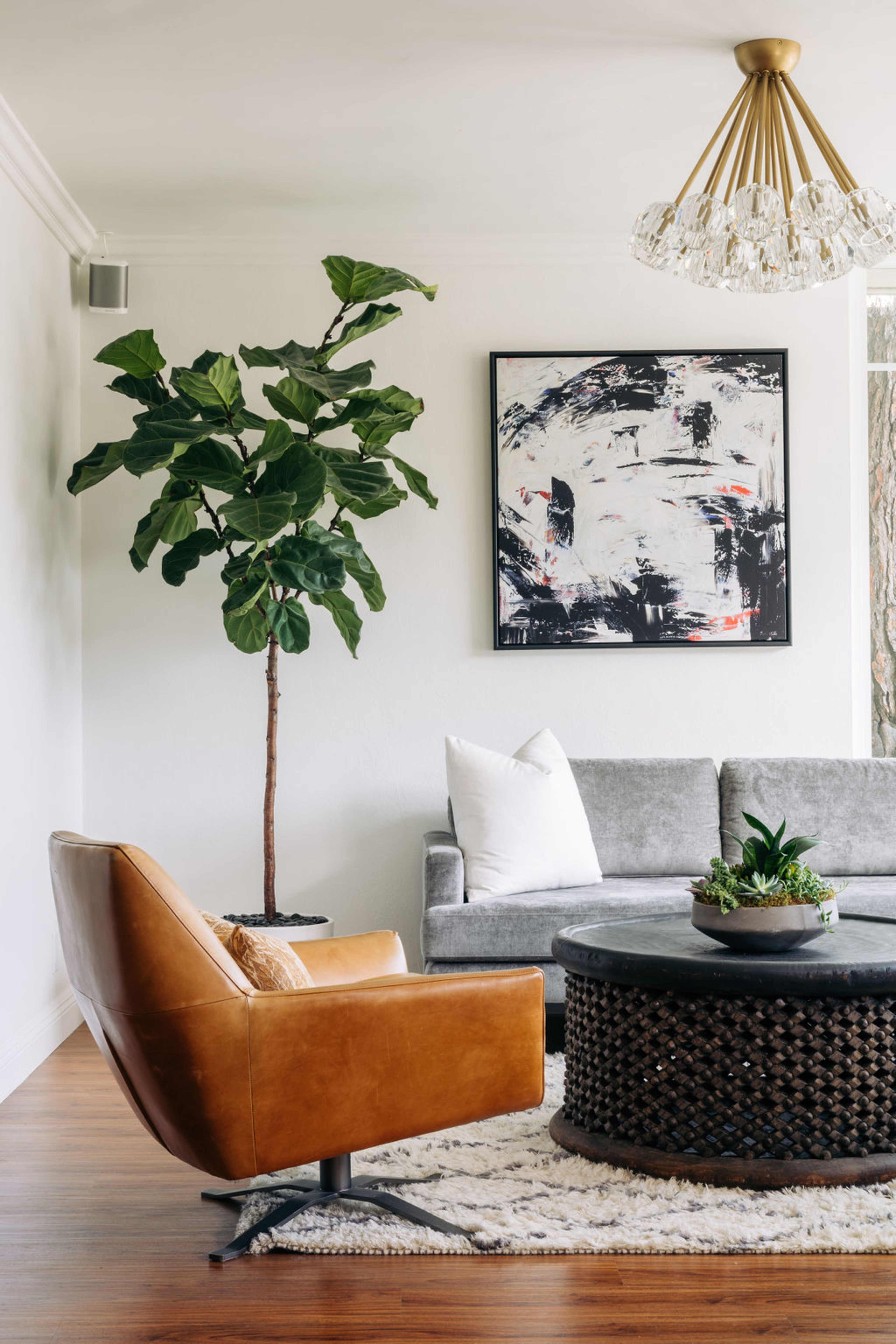 A modern living room featuring a gray sofa, a brown accent chair, a round coffee table, a large artwork on the wall, and a potted plant in the corner.