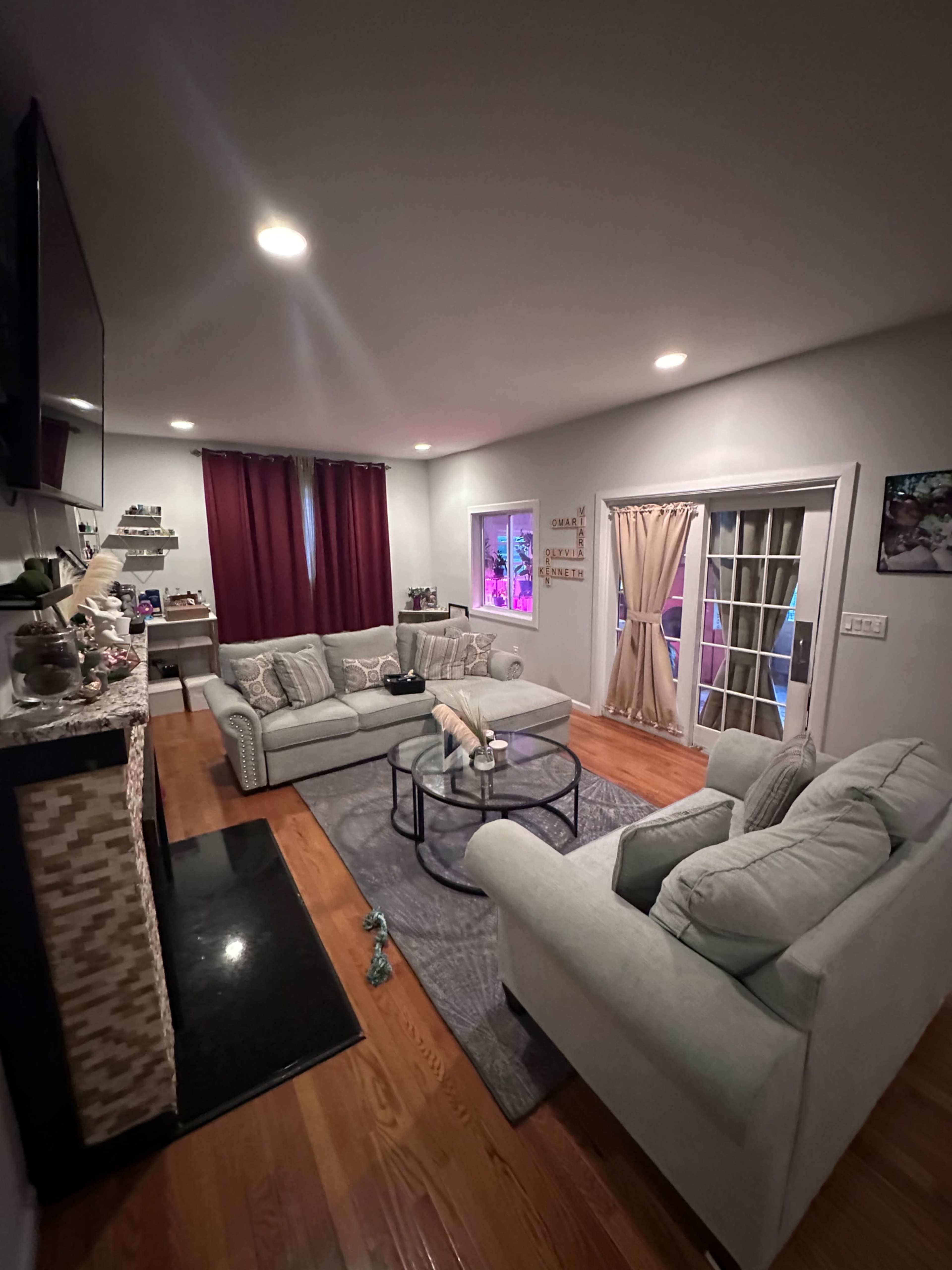 The image shows a cozy living room with a pair of light-colored sofas, a glass coffee table, and a fireplace, featuring dark curtains and a door leading to an outdoor area.