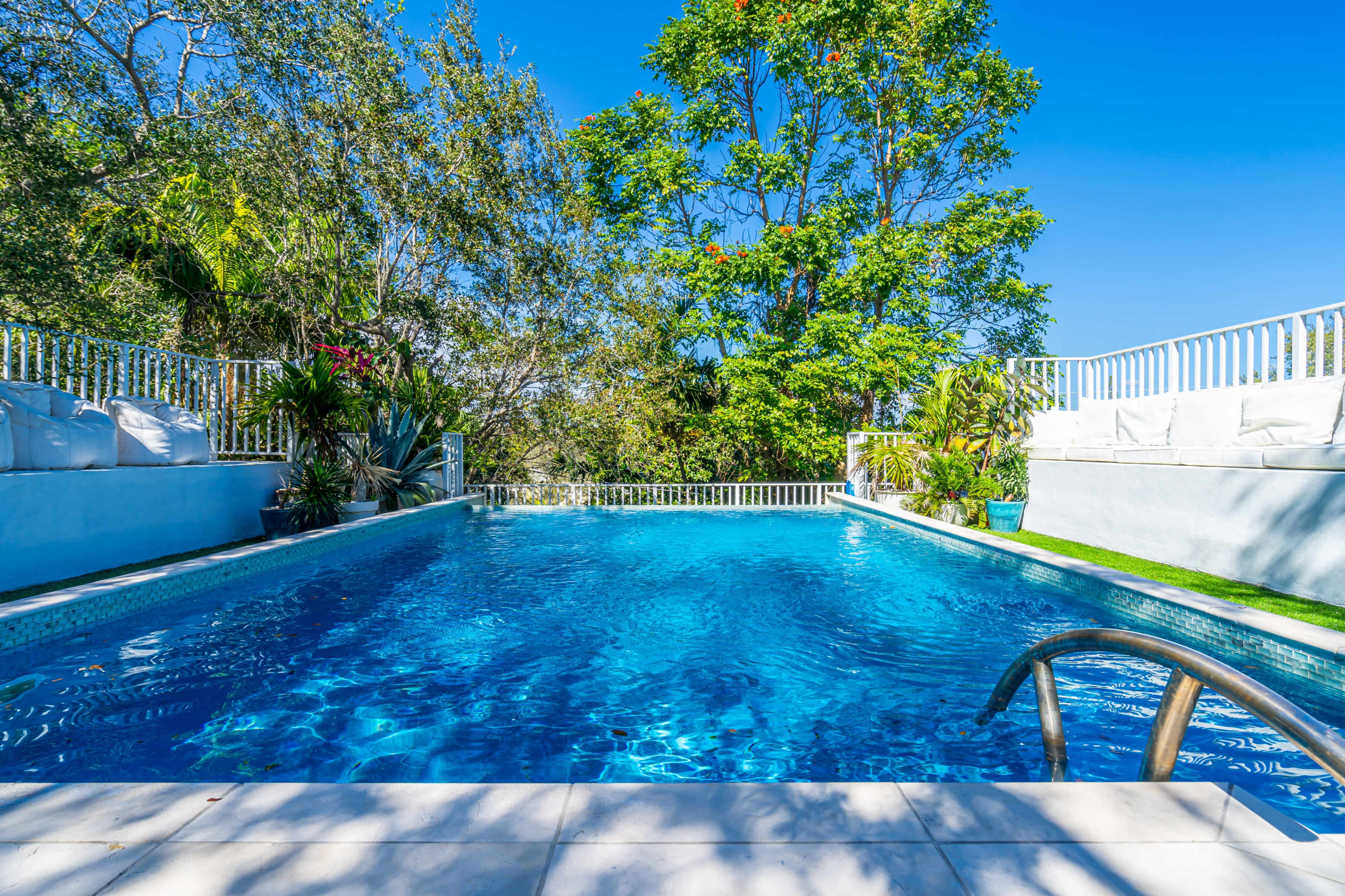 A clear blue swimming pool surrounded by lush greenery and white patio furniture under a blue sky.