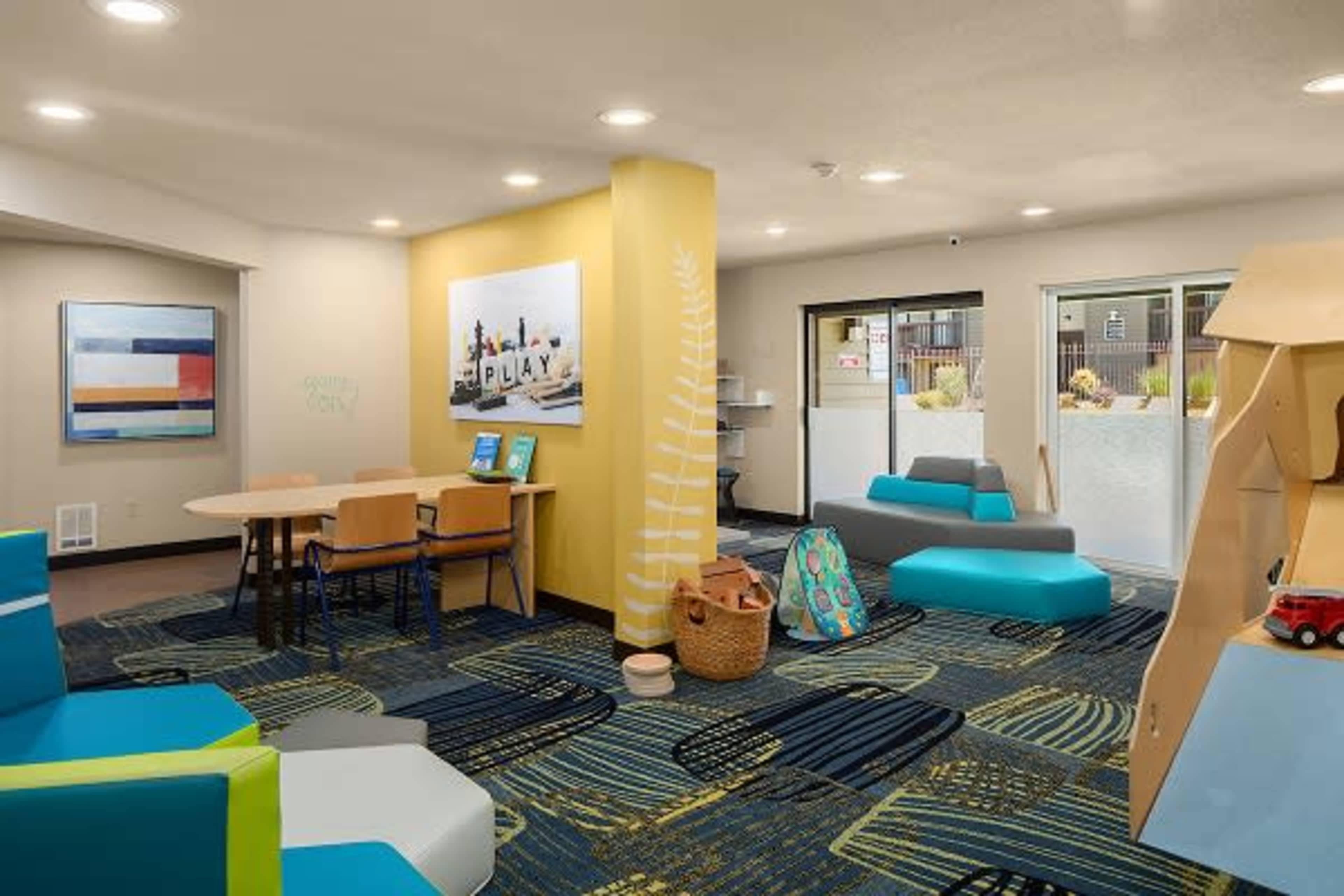 A colorful children's play area with seating, a table, and a view of an outdoor space through glass doors.