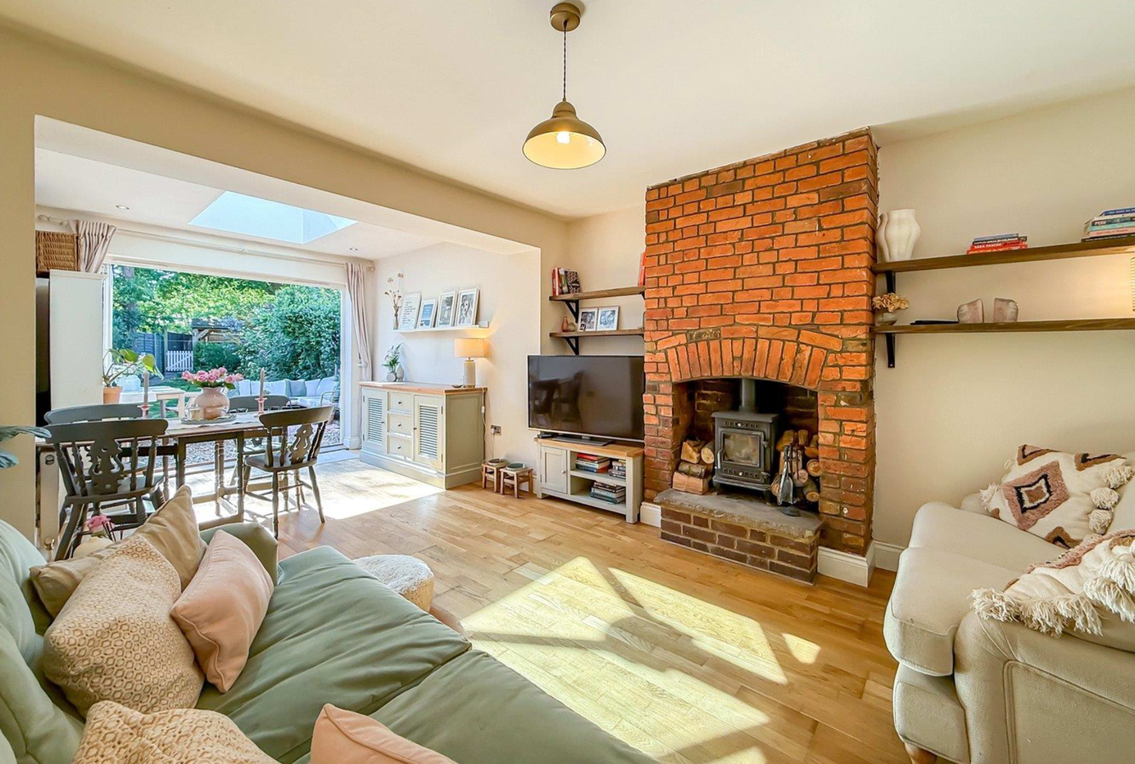 The image shows a cozy living room featuring a brick fireplace, a television mounted on the wall, and light wood flooring, with an open dining area visible in the background.