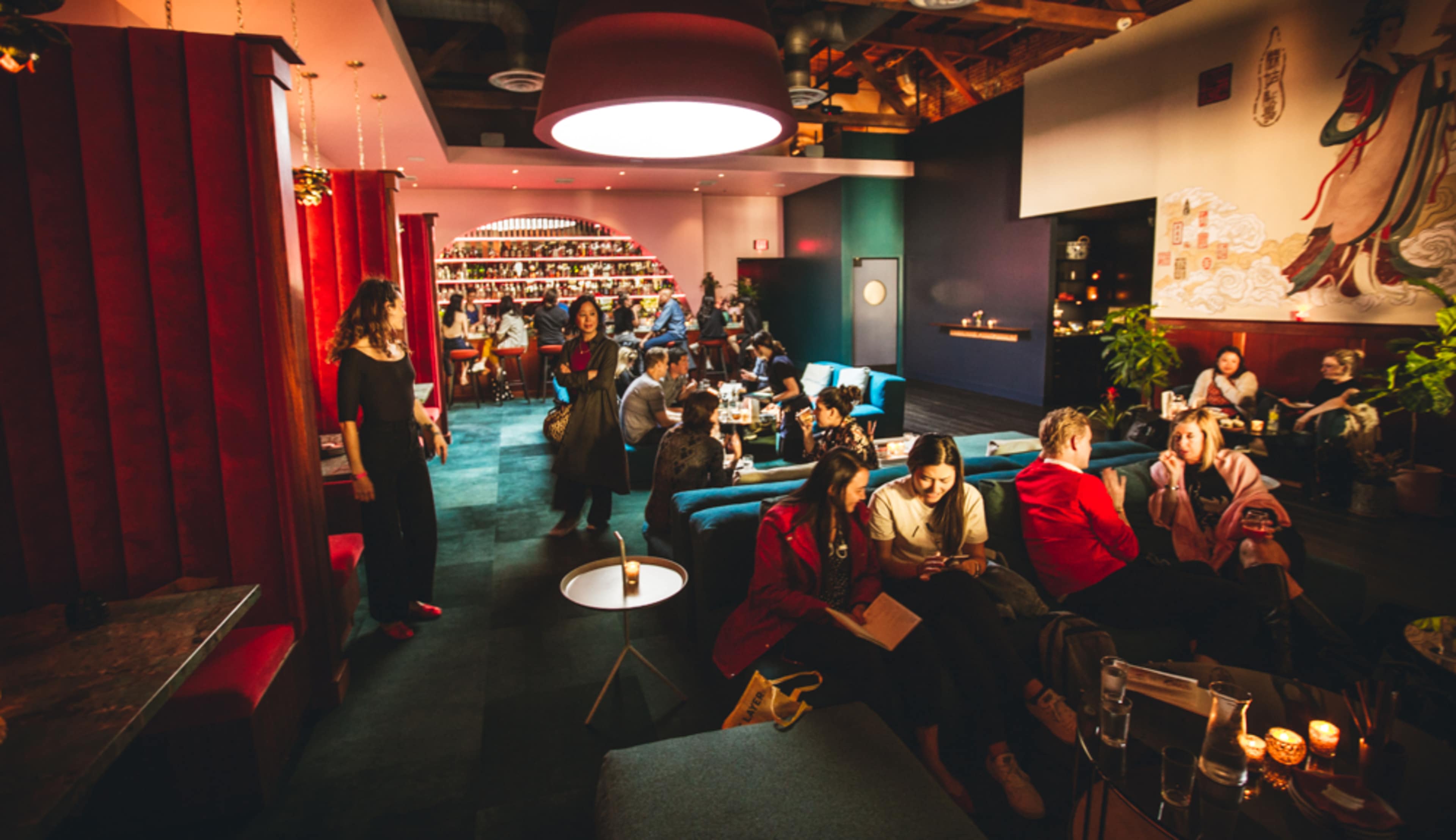 A lively lounge or café with people seated at various tables, some engaged in conversation and others looking at menus, surrounded by vibrant decor and a well-stocked bar.