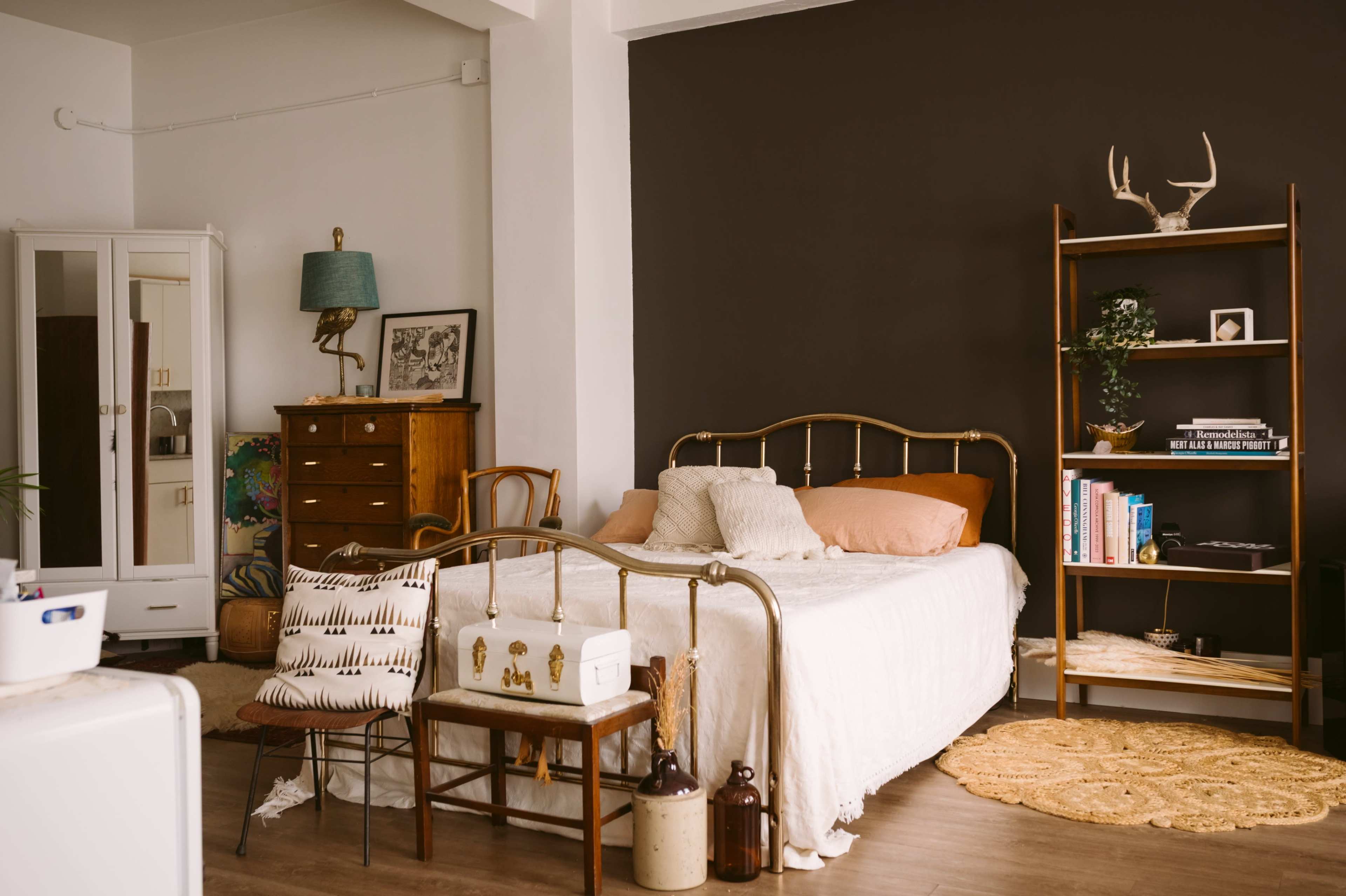 The image depicts a cozy bedroom featuring a brass bed frame, a wooden dresser, a bookshelf, and decorative elements like a lamp and plants against a dark accent wall.