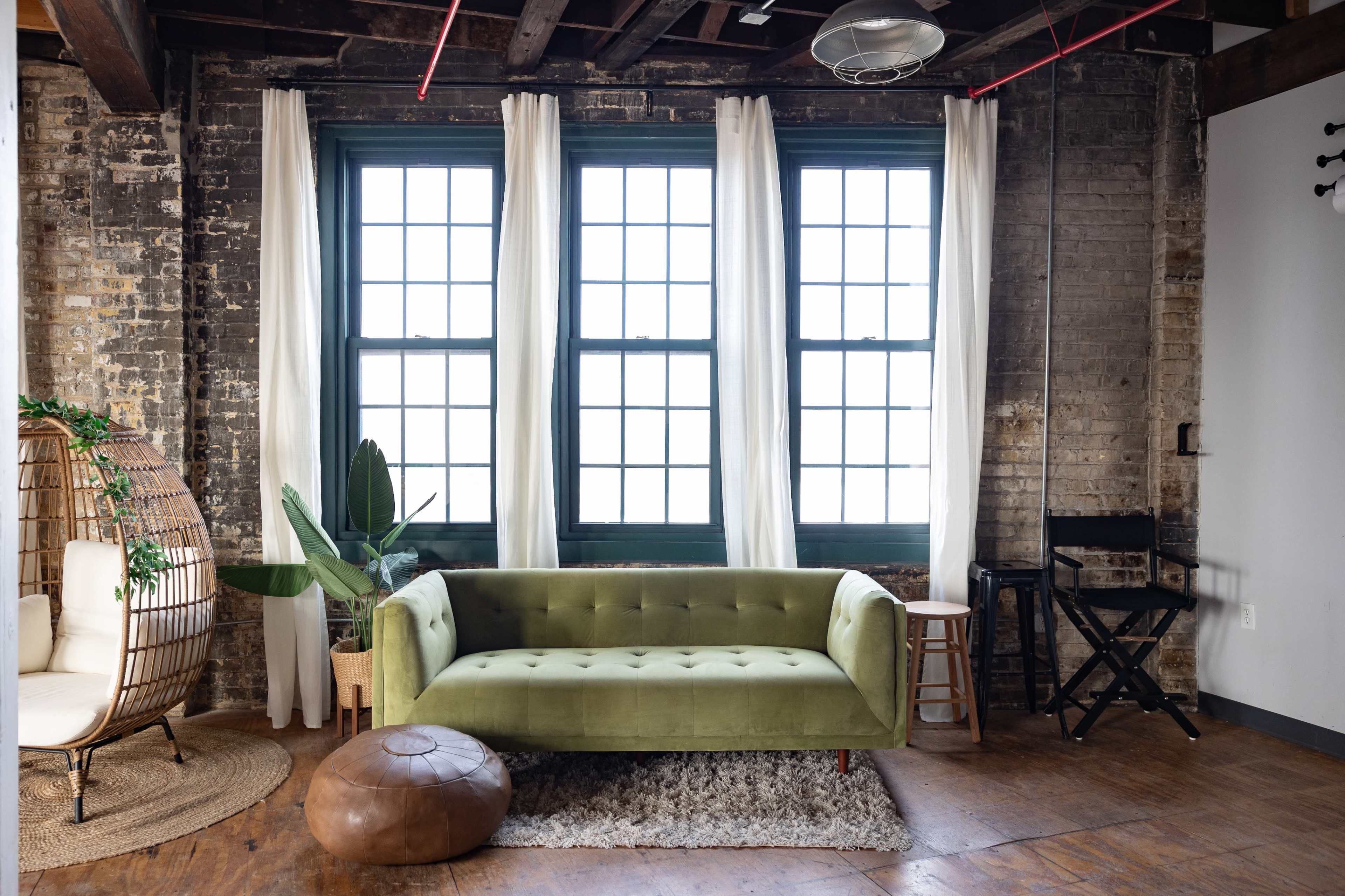 The image shows a cozy living space with a green sofa, large windows, sheer curtains, a potted plant, and a round pouf on a textured rug.