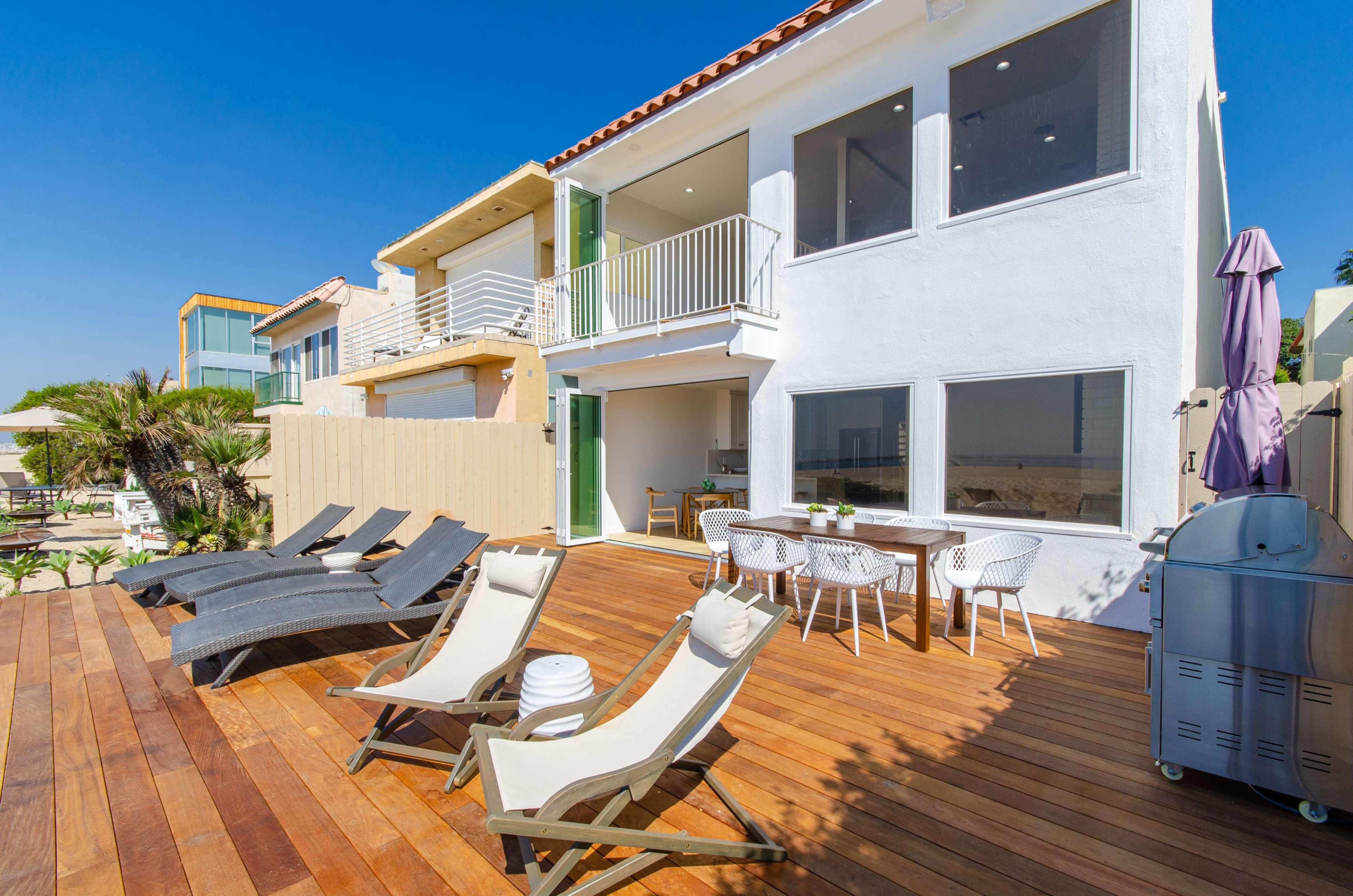 A modern beach house with a spacious wooden deck featuring lounge chairs and an outdoor dining area.