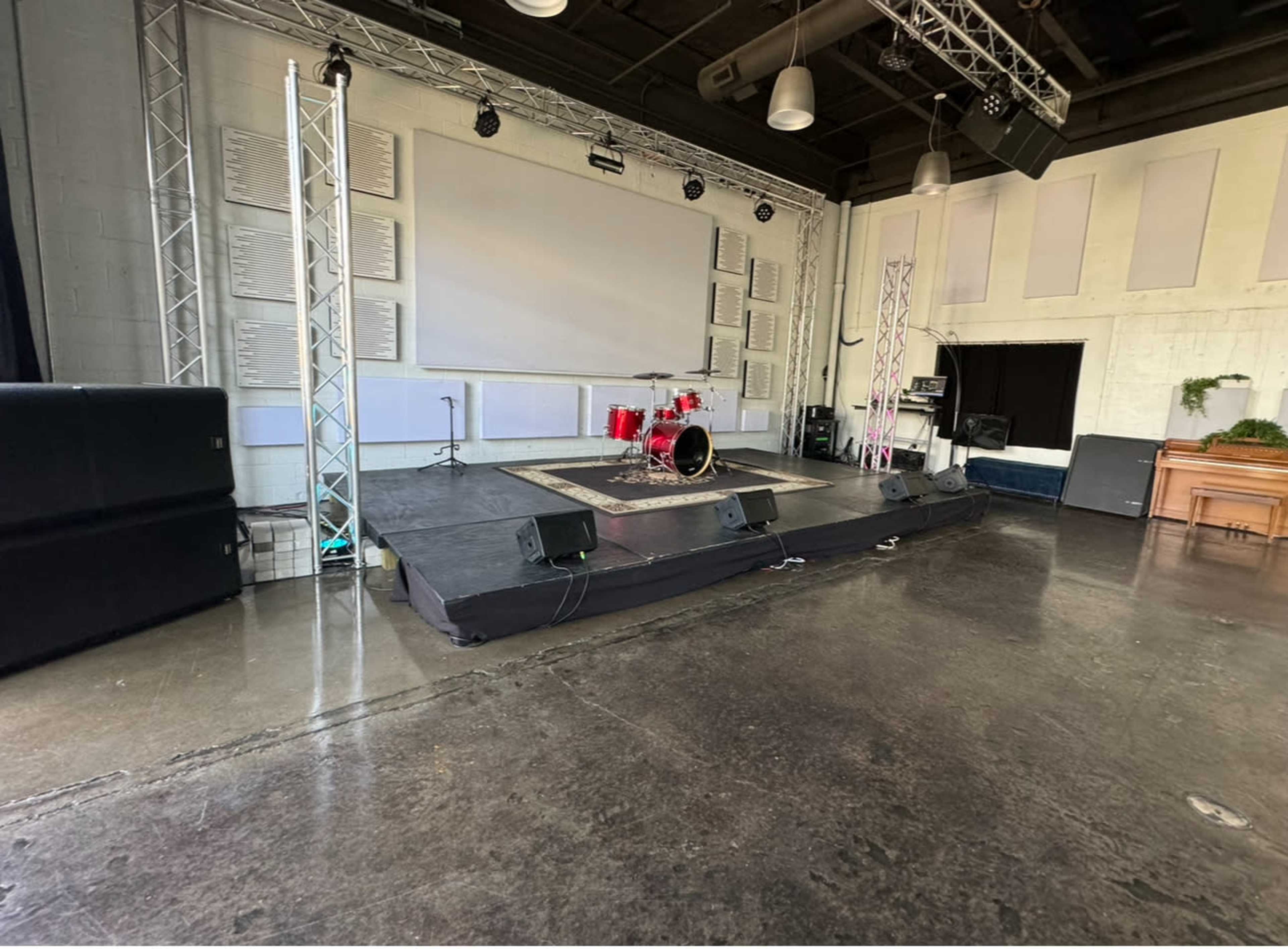 The image shows a stage setup in a spacious room, featuring a drum kit and sound equipment against a backdrop of a large screen.