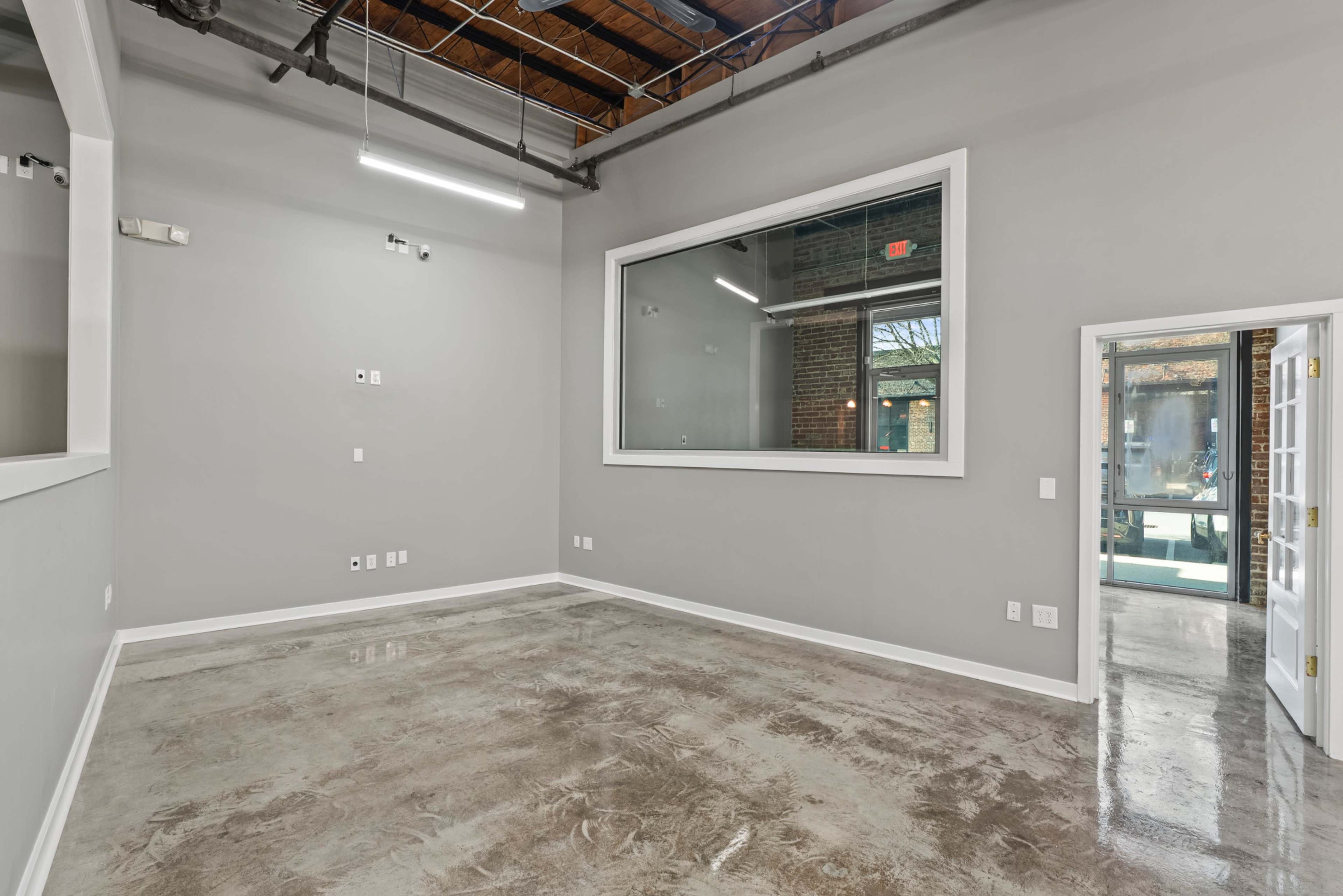 The image shows a spacious, empty room with polished concrete floors, gray walls, and a large windowed area on one side.