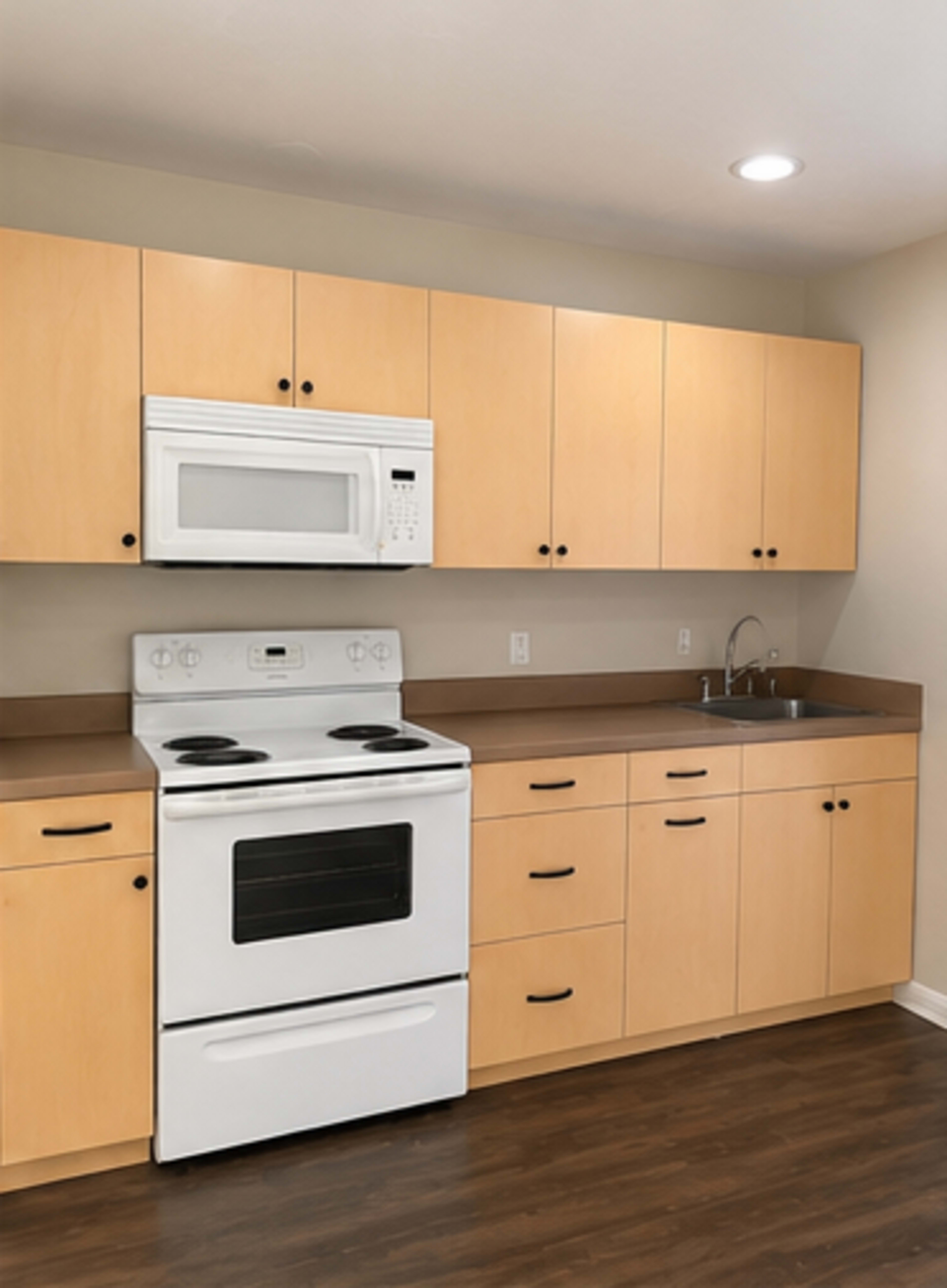 The image shows a functional kitchen equipped with light wood cabinetry, a white stove, a microwave, and a sink.
