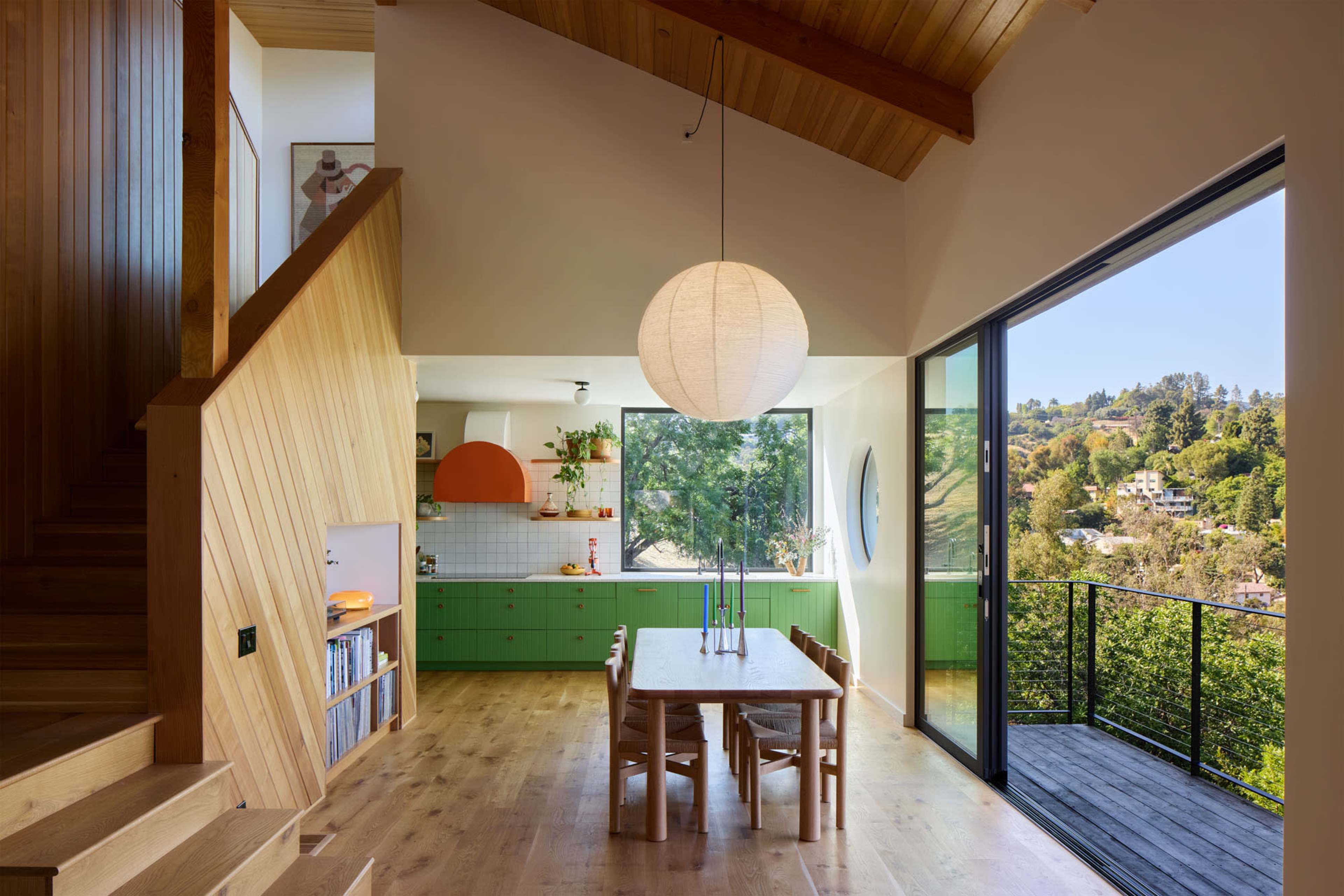 The image shows a modern interior of a house featuring a wooden staircase, a green kitchen with a round orange accent, and a dining area with a wooden table and a large window that opens to a view of a hilly landscape.