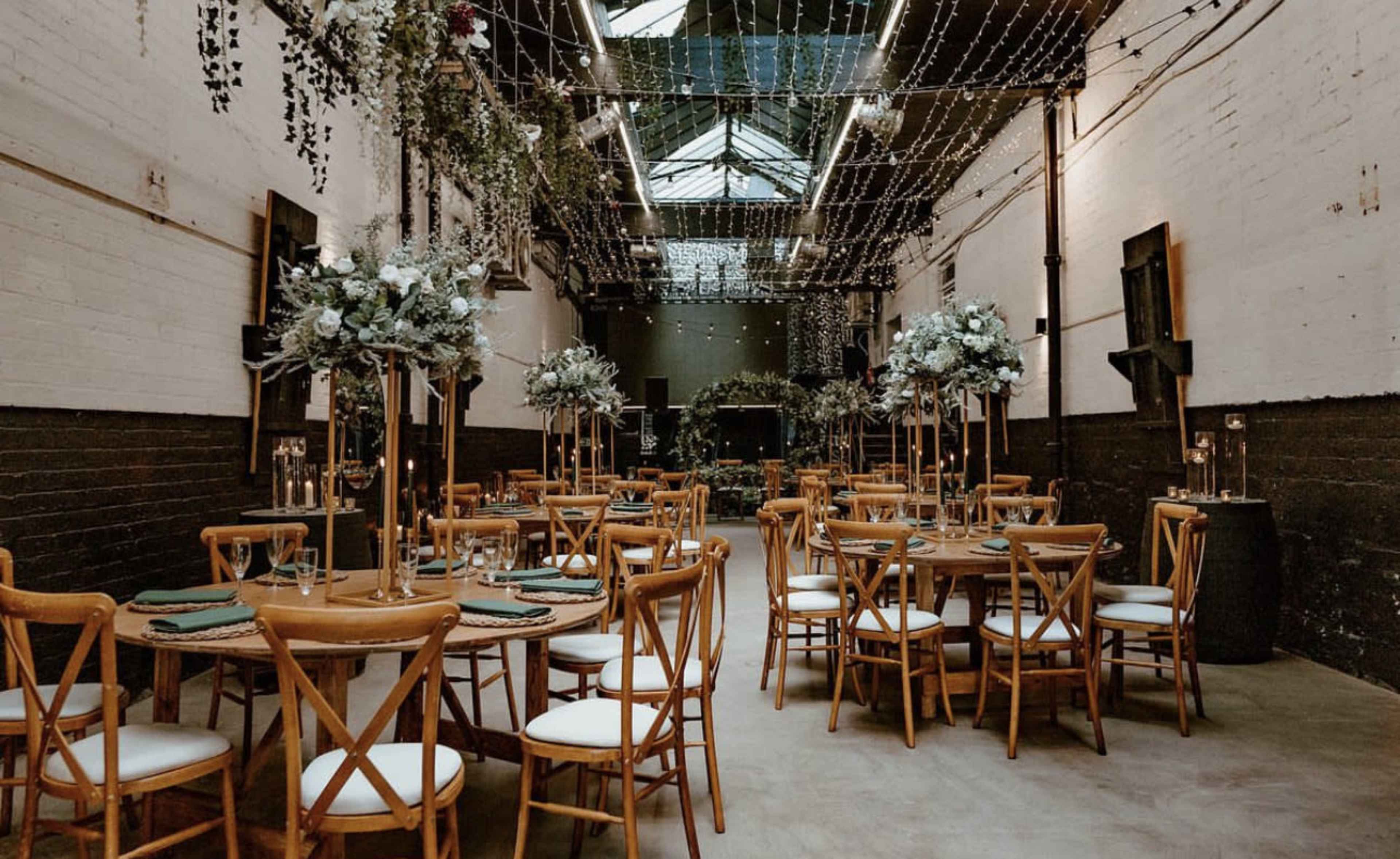 A spacious, elegantly arranged dining area features wooden tables with white chairs, surrounded by floral centerpieces, under soft string lights and a skylight.