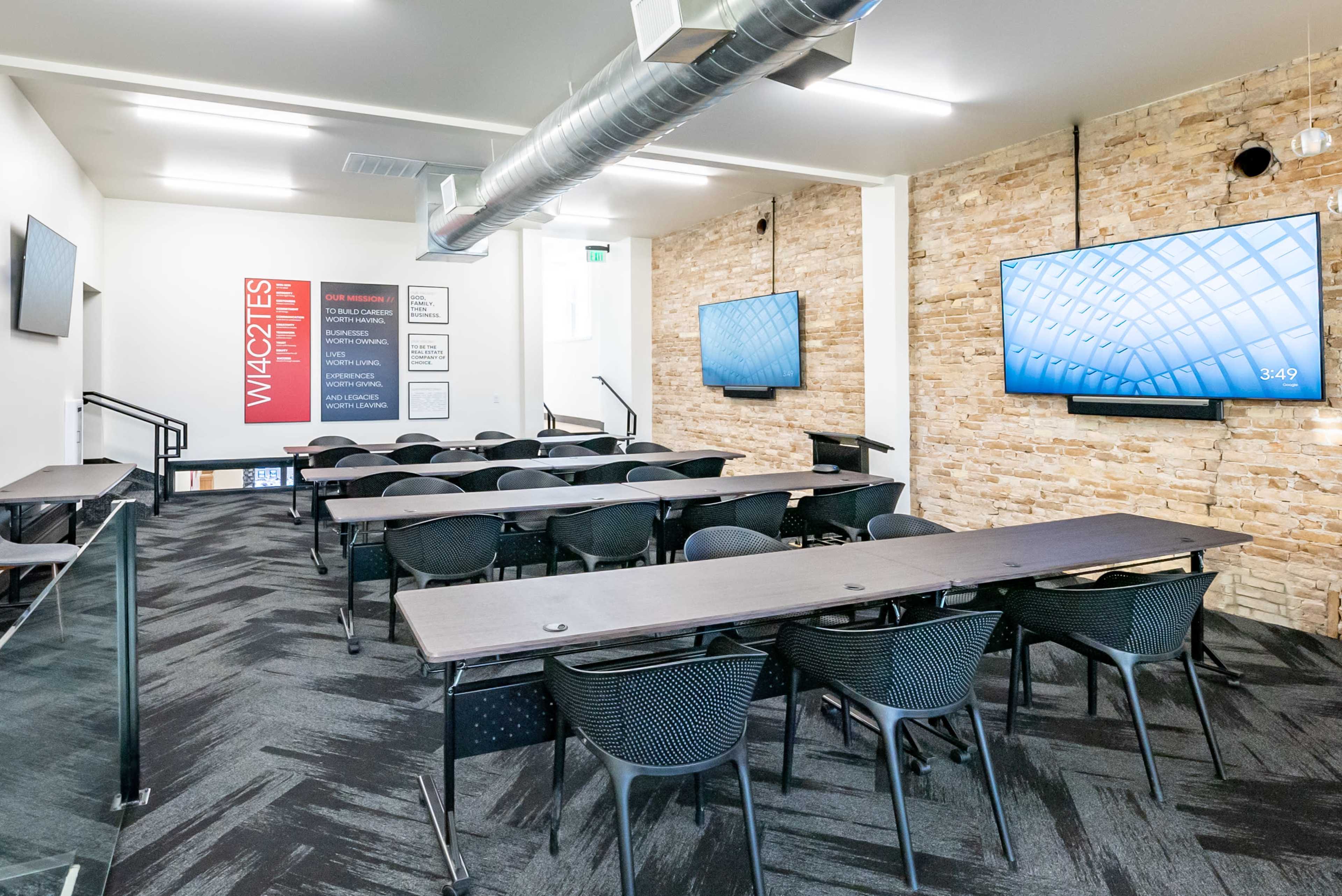 A modern classroom with rows of black chairs and tables, equipped with two large screens mounted on the walls and exposed stone accents.