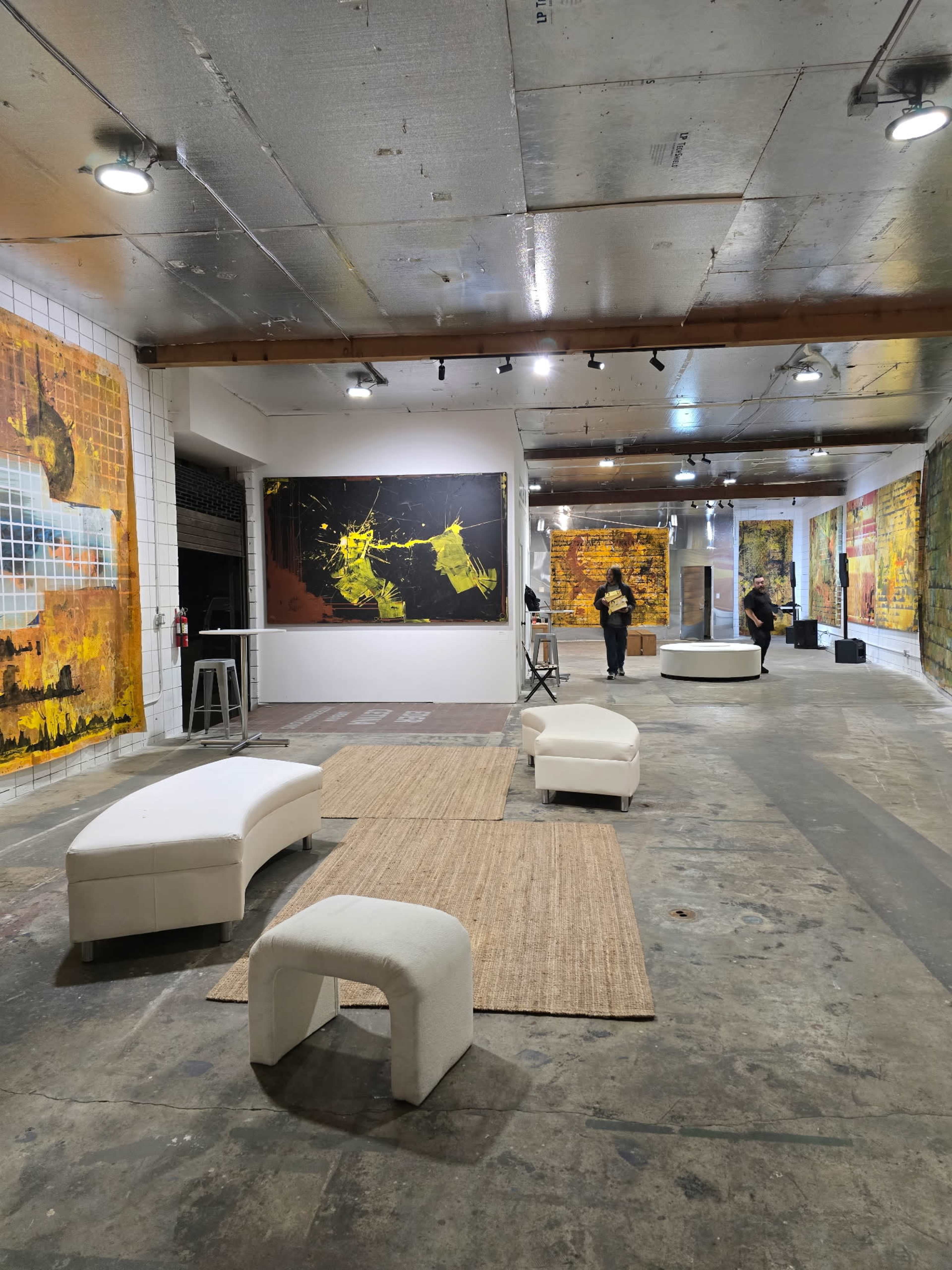 The image shows an art gallery featuring large abstract paintings on the walls, with seating areas and visitors engaging with the artwork.