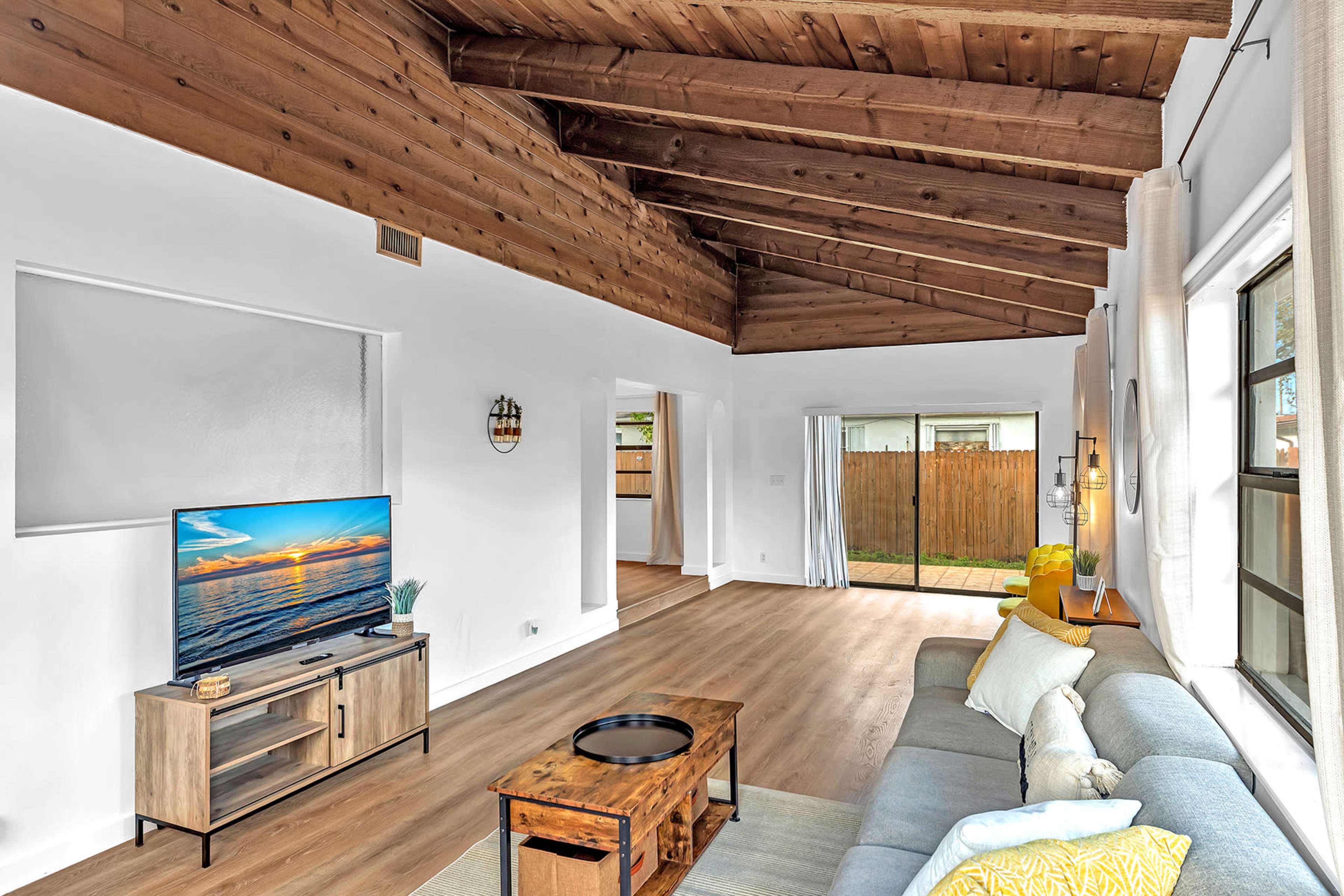 A spacious living room features a wooden beam ceiling, a large TV on a stand, a gray sofa, and sliding glass doors leading to a backyard.