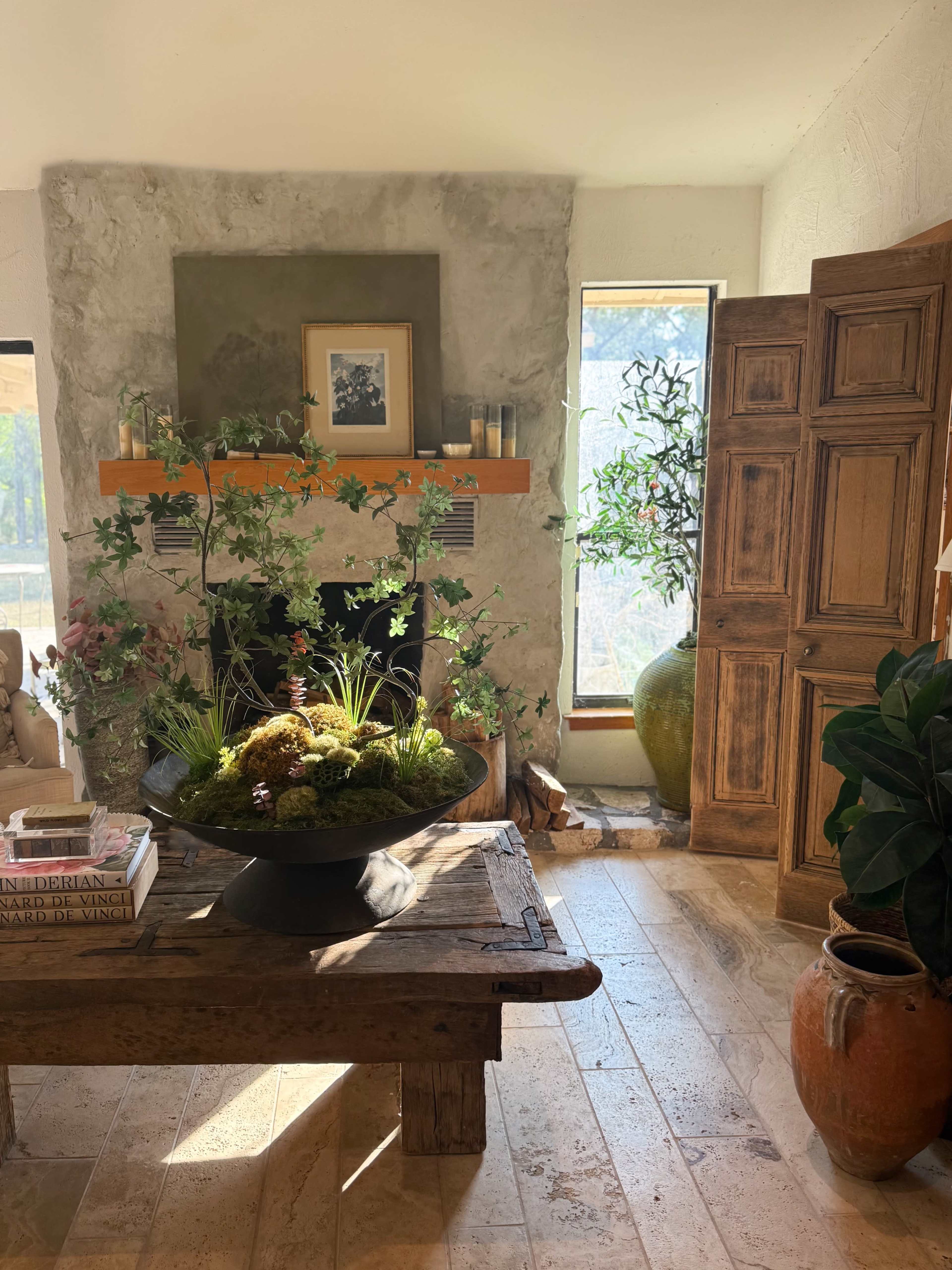 A cozy interior features a wooden coffee table with decorative books, a large bowl of plants, and natural light streaming through a window, complemented by rustic wooden cabinets and ornamental vases.