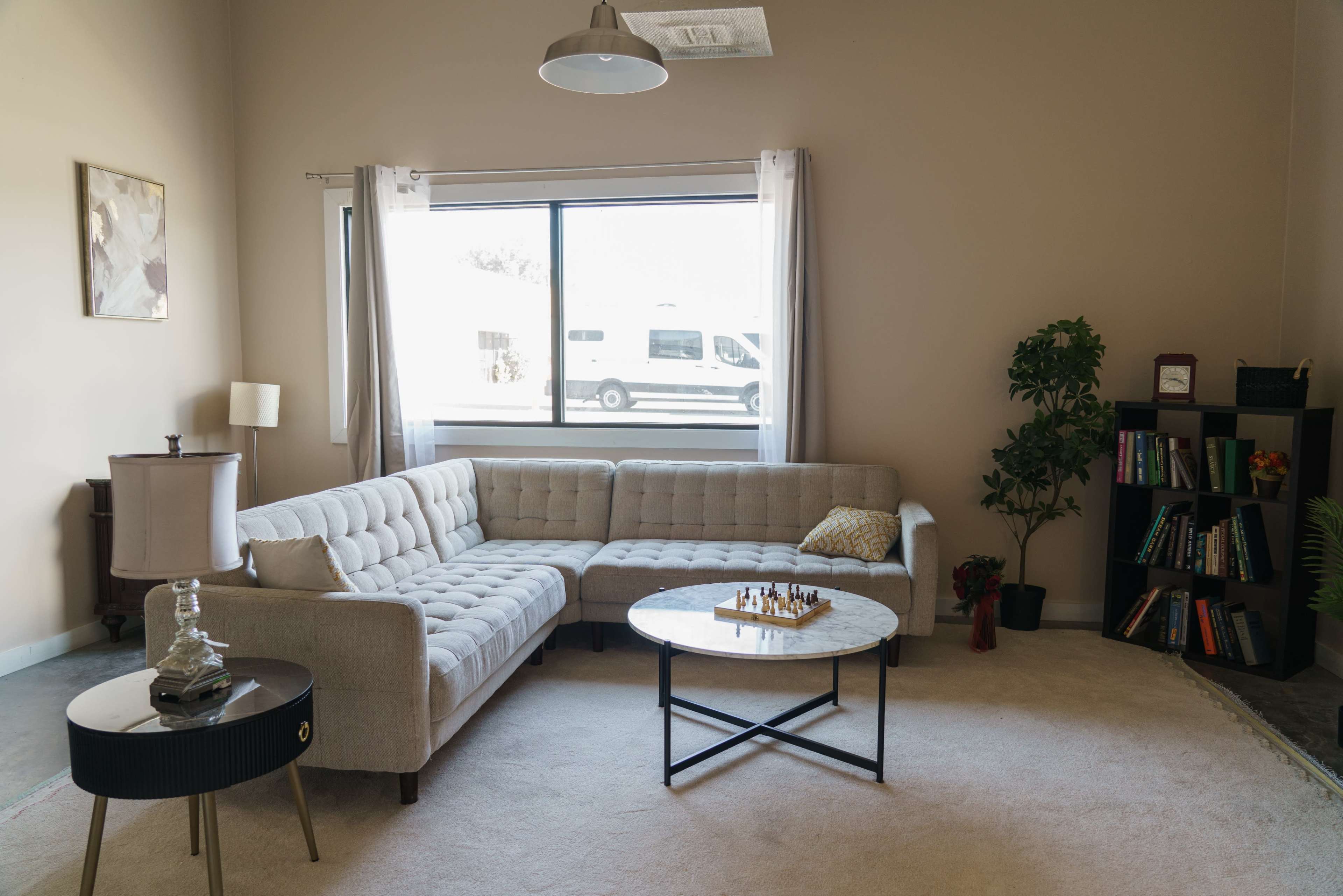 A modern living room features a beige sectional sofa, a round coffee table with a chess set, and a window exposing an outdoor view, complemented by a small plant and a bookshelf.