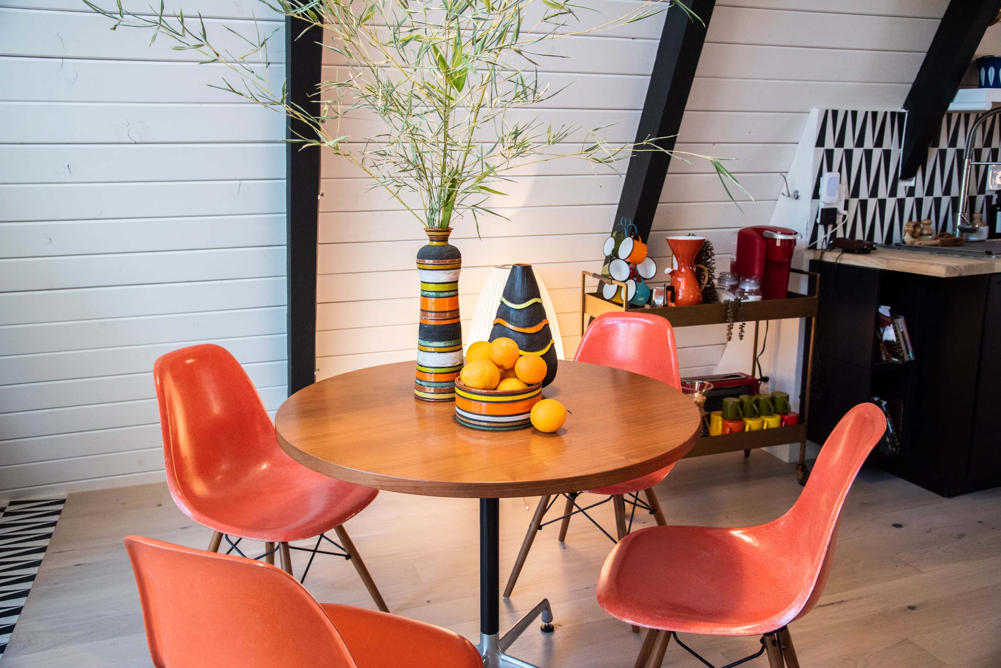 A round wooden dining table is set with a bowl of oranges and surrounded by four red chairs, with decorative vases and a kitchen area visible in the background.