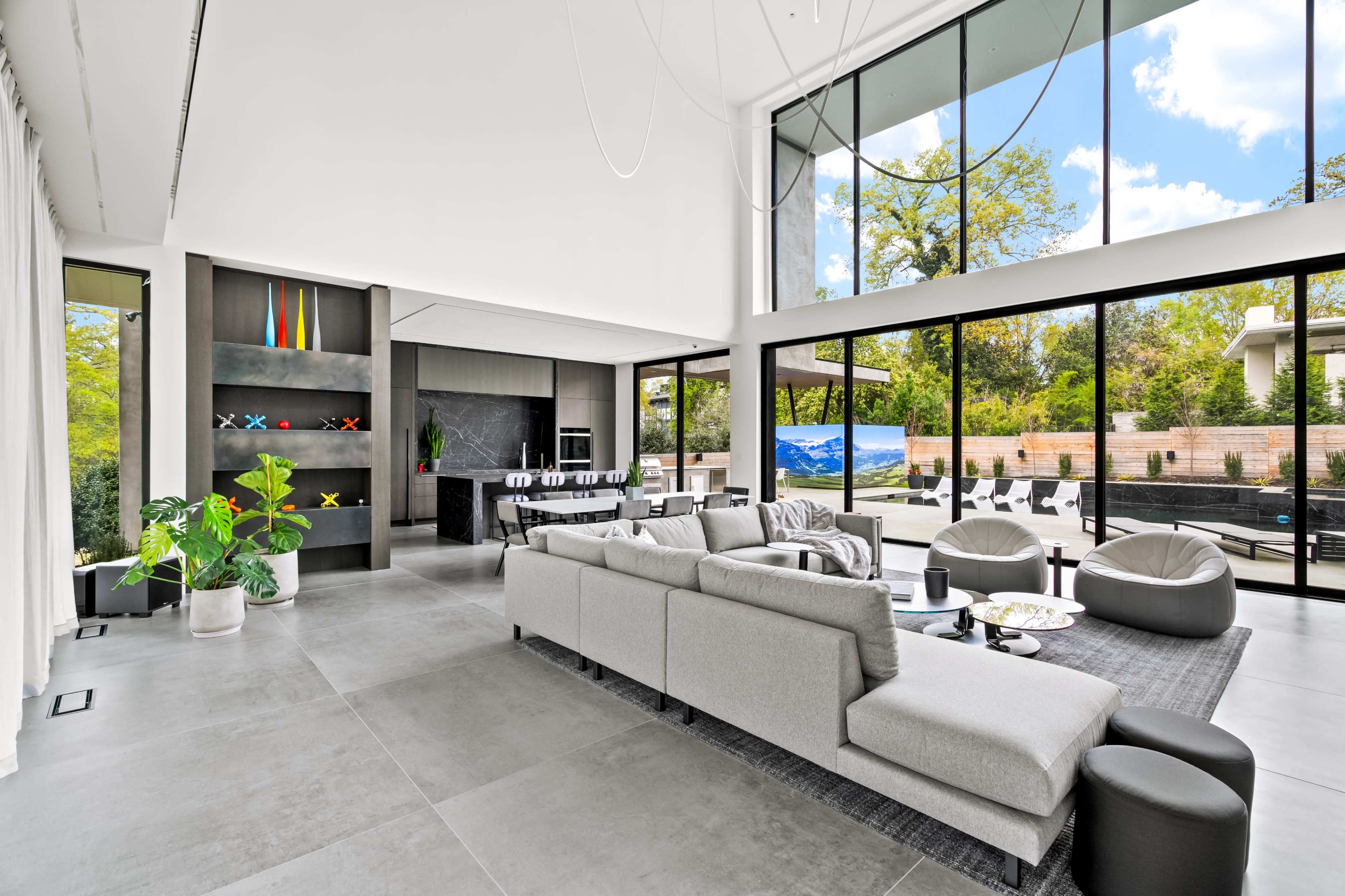 The image shows a modern living room with large windows, a sectional sofa, a minimalist design, and an open layout that leads to an outdoor pool area.