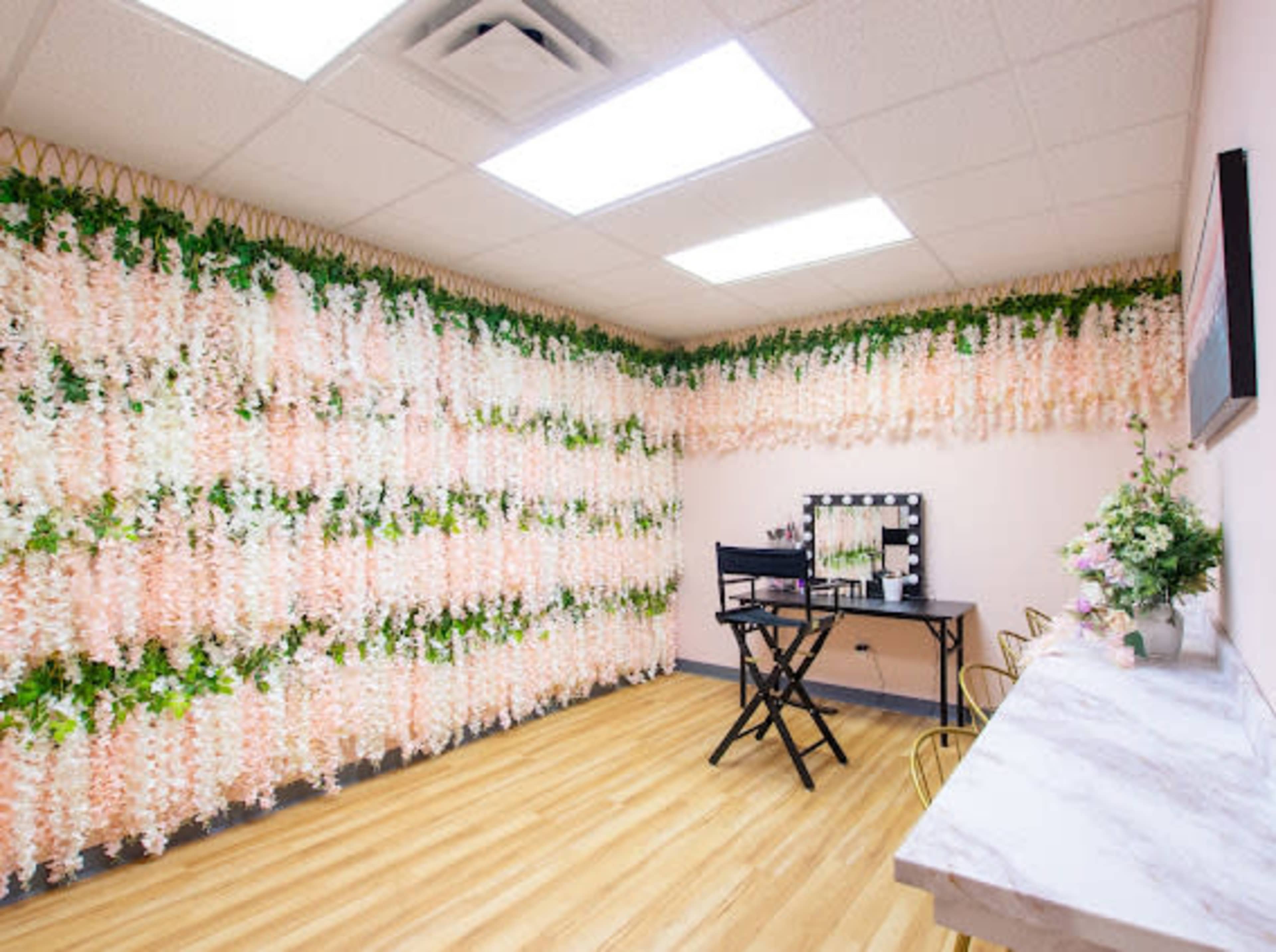 A bright room features a floral wall of white and pink flowers, a black makeup chair, a mirror with lights, and a marble countertop.