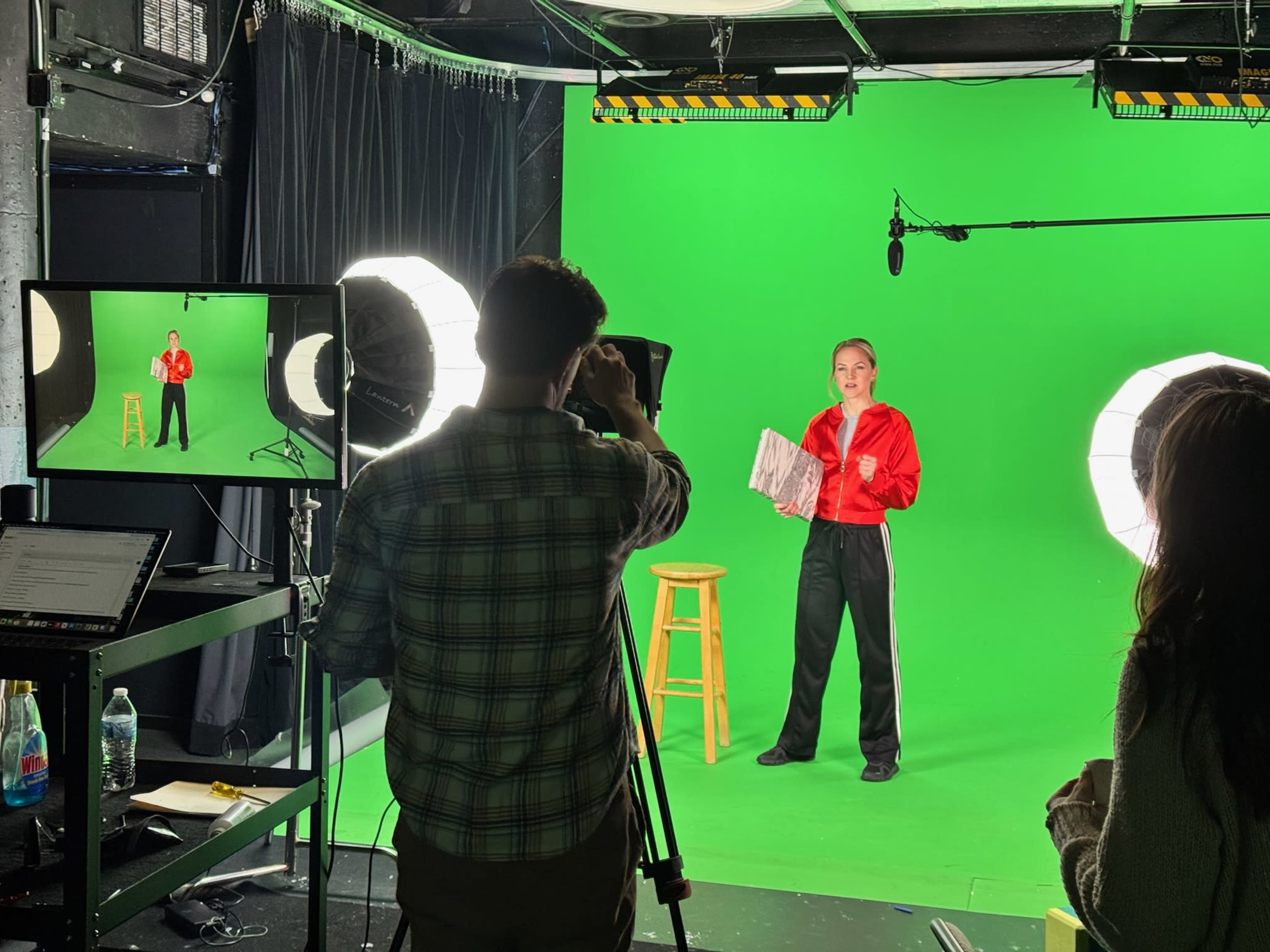 A person in a red jacket stands on a green screen stage holding a sheet of paper while being filmed by a cameraman.